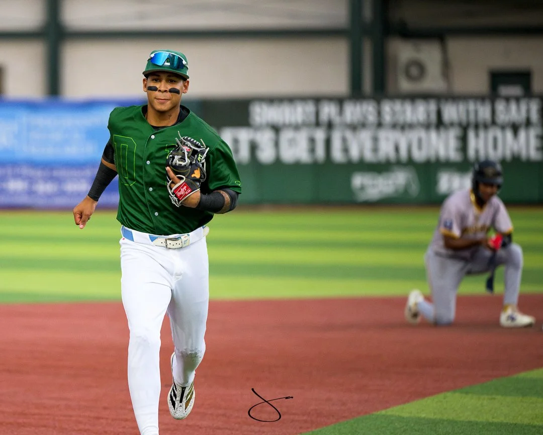 Baseball player running on the field in green and white uniform, wearing a helmet and holding a baseball glove, with another player kneeling in the background.