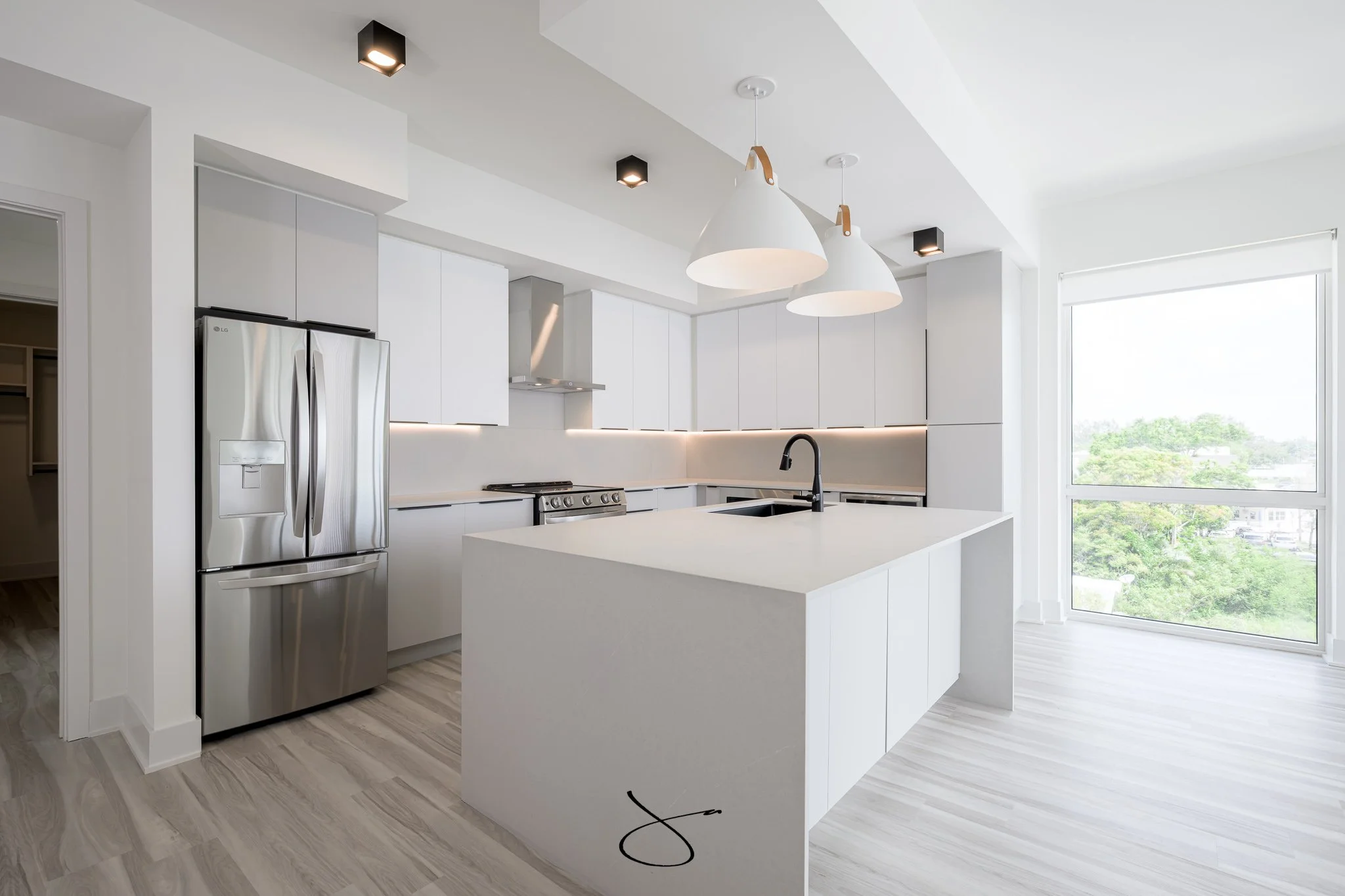 A modern, minimalist kitchen with white cabinets, a central island, stainless steel refrigerator, and a large window letting in natural light.