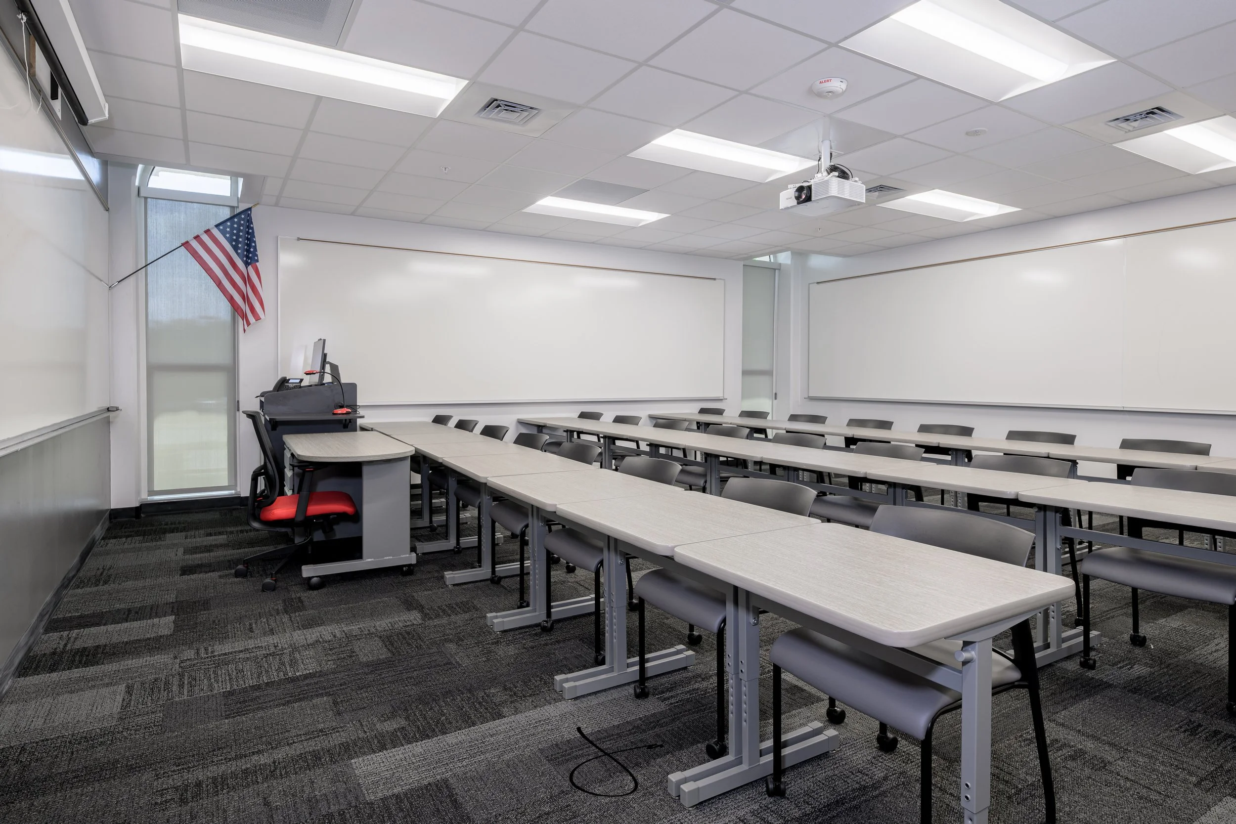 An empty classroom with white walls, a row of tables and chairs, a whiteboard, and a mounted projector on the ceiling. There is an American flag hanging near the window.