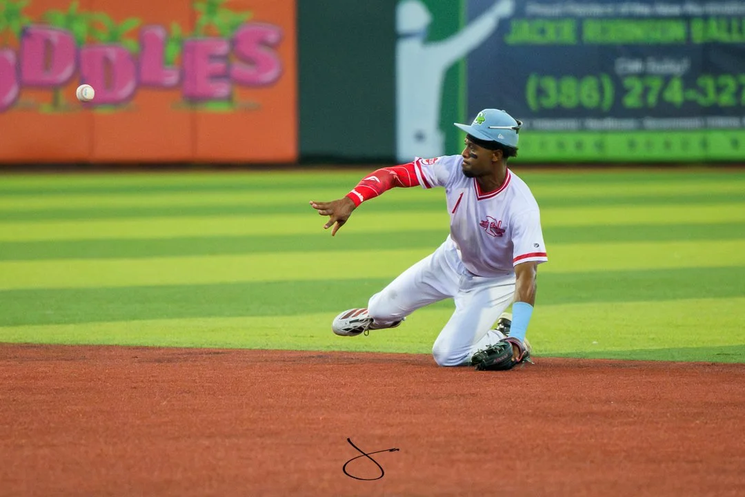A baseball player is kneeling on the ground and reaching for a ball that is mid-air. The player is wearing a white uniform with red accents, a batting helmet, and a protective arm sleeve. The field has bright green grass and reddish-brown infield dir