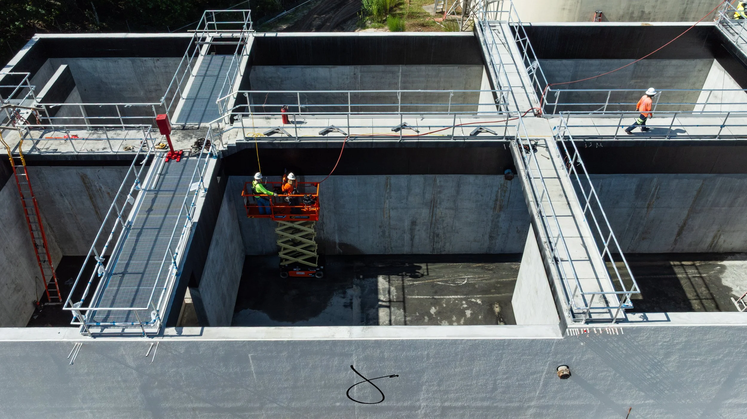 Workers in safety gear installing or inspecting large concrete water treatment tanks with metal walkways and safety railings at a construction site.