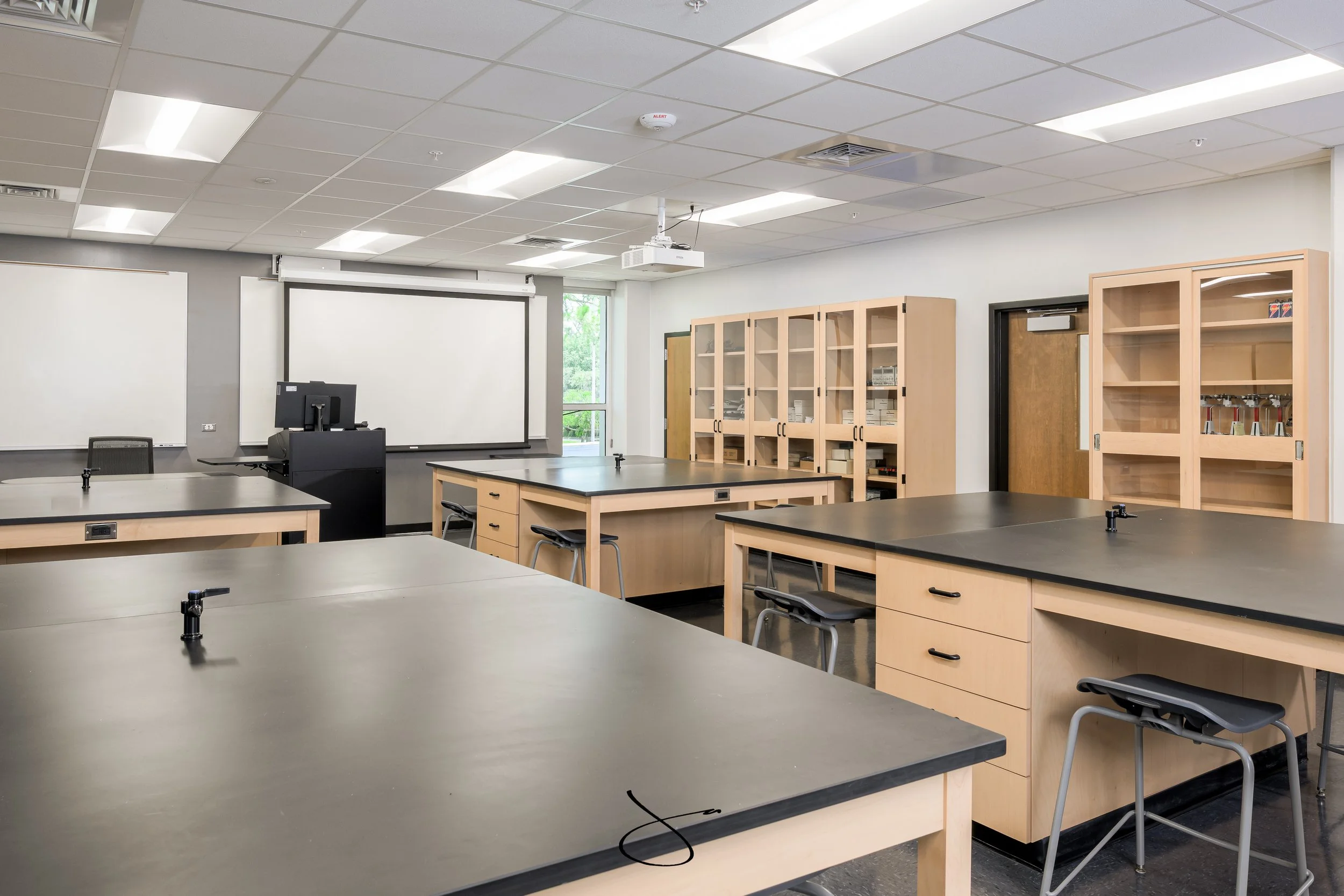 Empty classroom with desks, chairs, a computer, cabinets, whiteboards, and a ceiling-mounted projector.