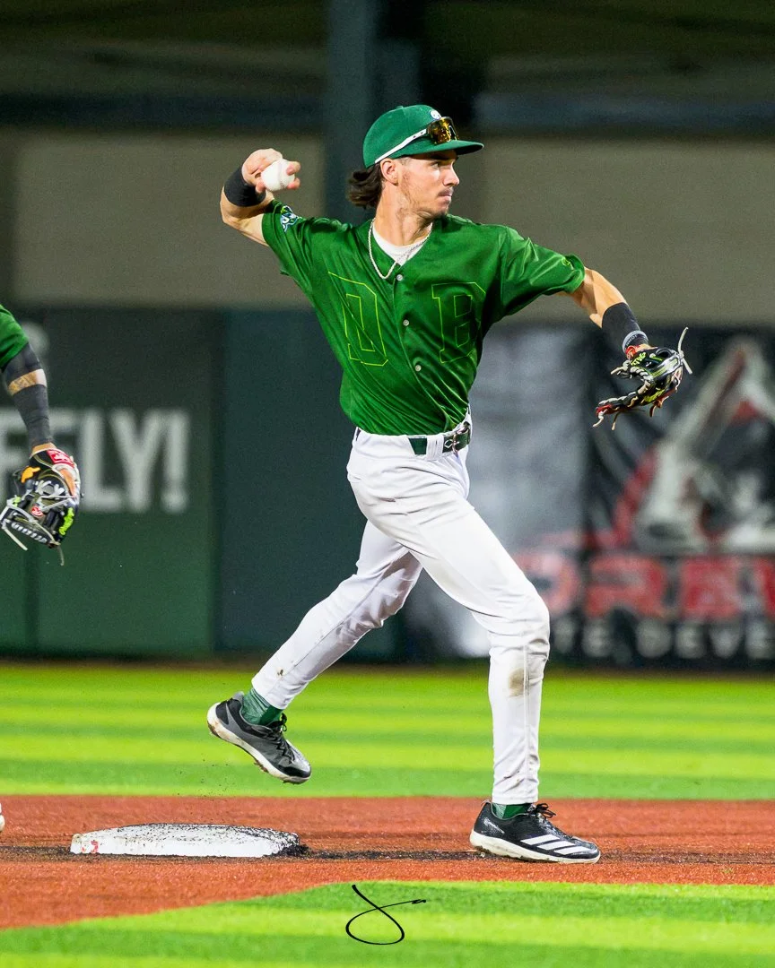 A baseball player in a green uniform and cap is running the bases, holding a baseball in his right hand and wearing black and white cleats on a well-lit field.