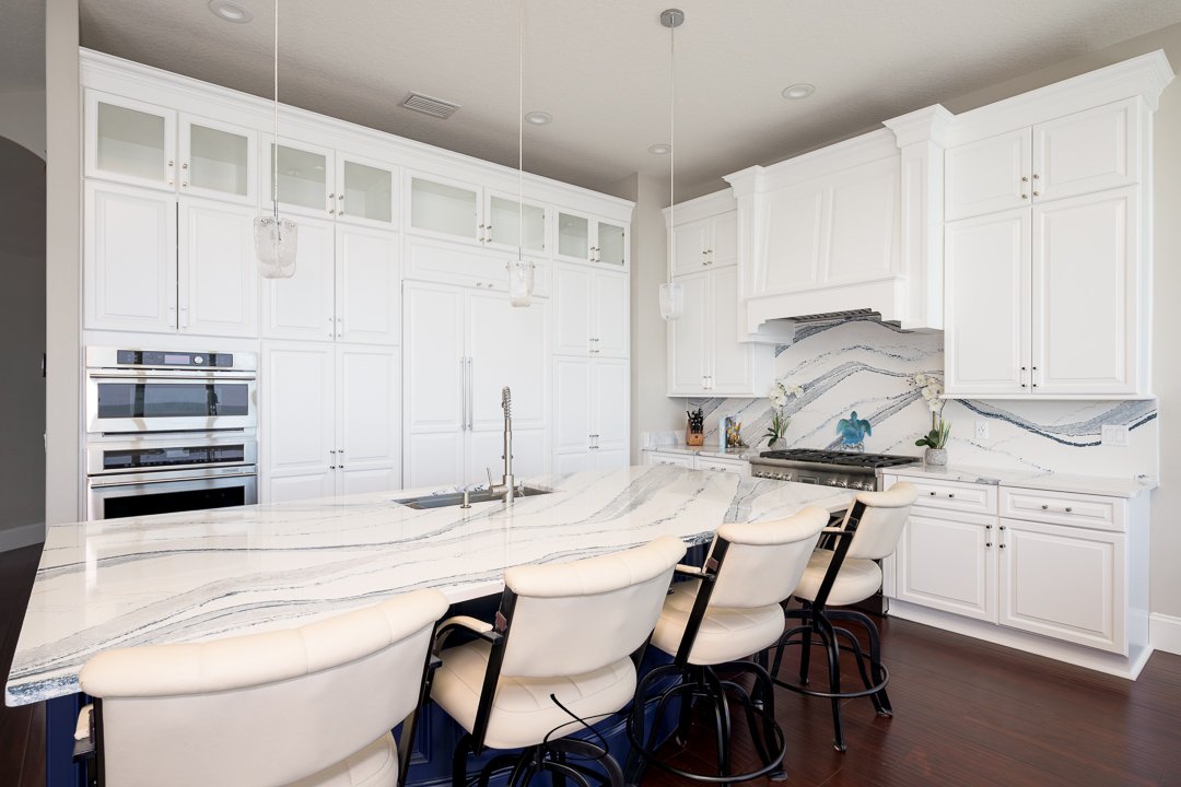 Modern white kitchen with marble countertops and backsplash, white cabinetry, and island with four white chairs.