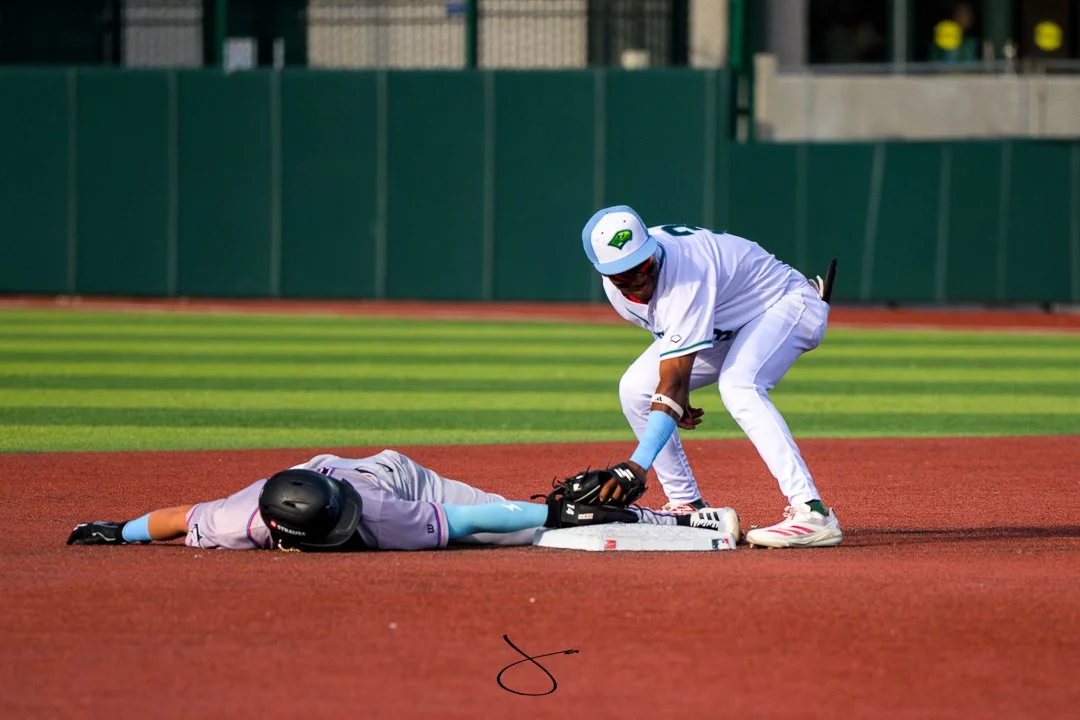 A baseball player lying face down on the infield dirt, with another player assisting him by placing a foot on his back near second base.