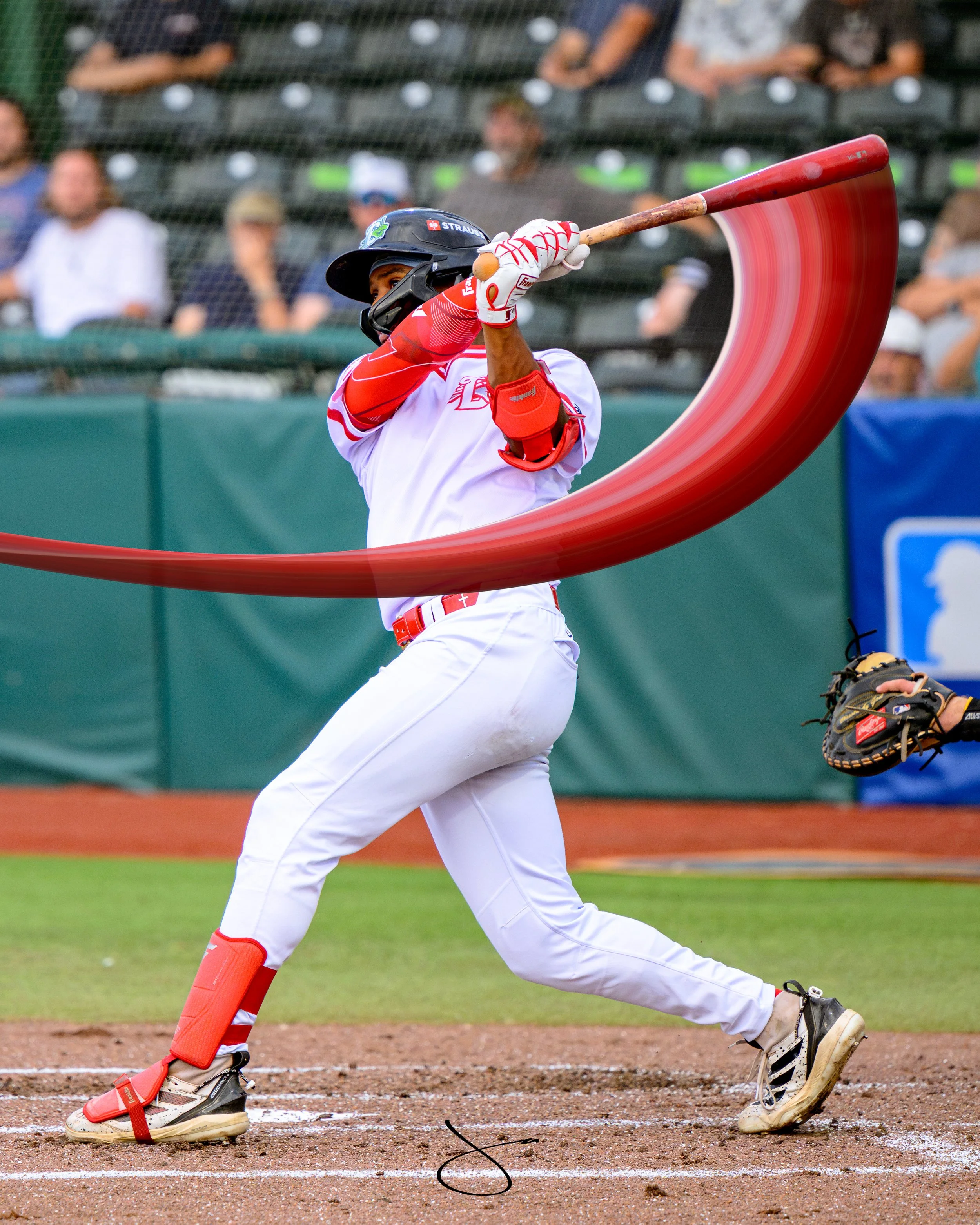 Female baseball player swinging a bat at the ball, wearing a black helmet, red and white uniform, and protective gear on her legs at a baseball field with spectators in the background.