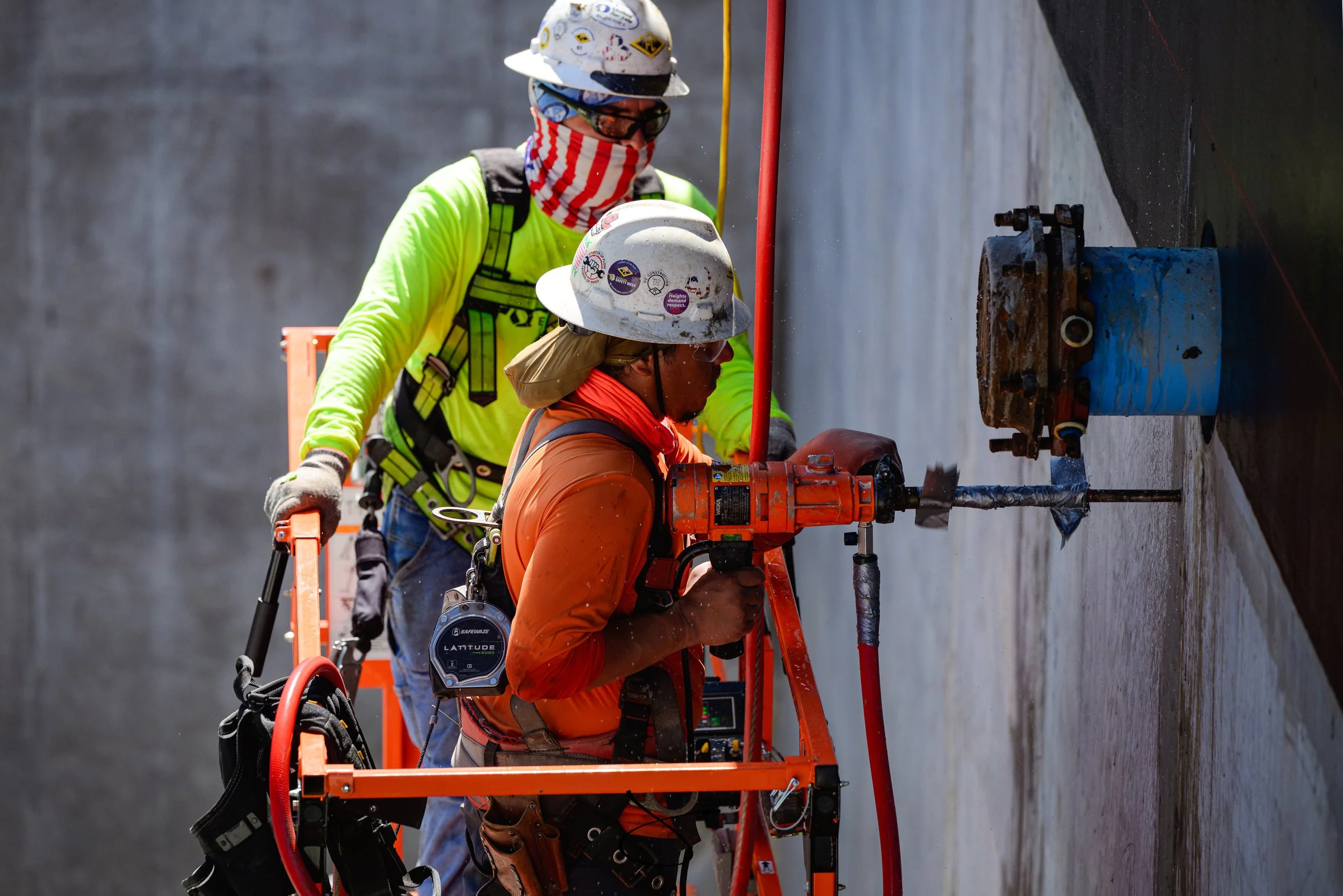Two construction workers wearing safety helmets, glasses, and bright safety clothing working on an industrial pipe or mechanism on a vertical wall.