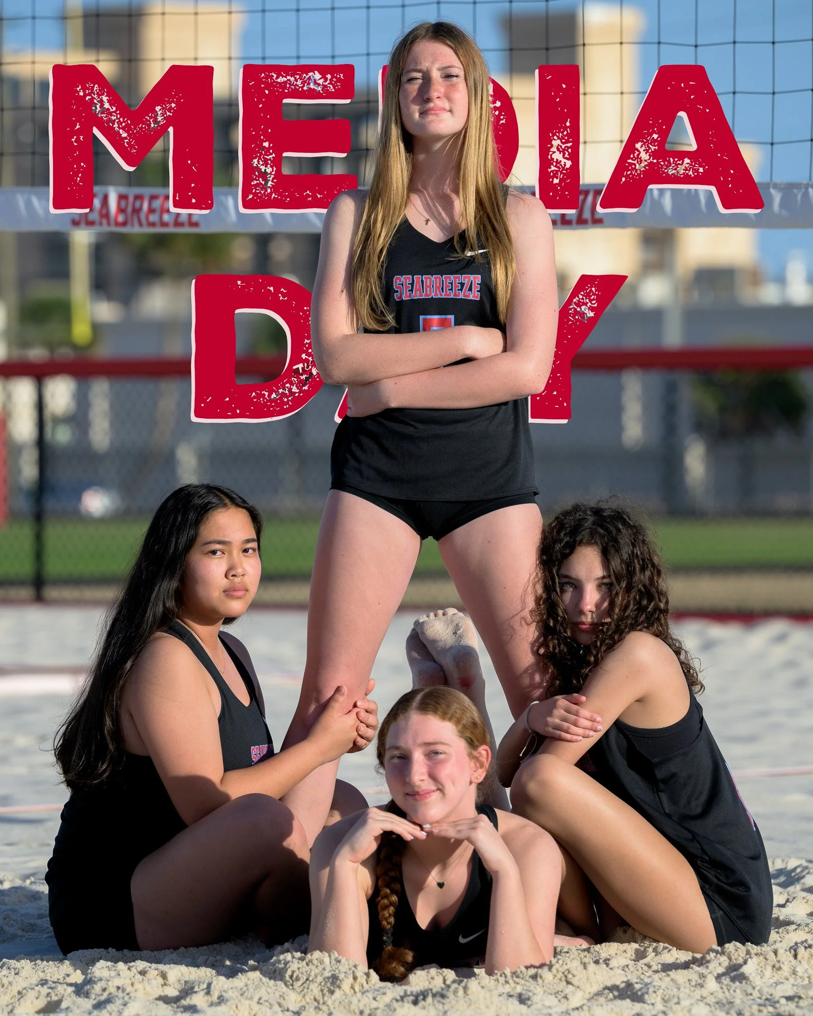 Group of four young women in athletic wear on a beach volleyball court, with one standing in the center and two sitting on the sand, and one lying down, posing confidently.