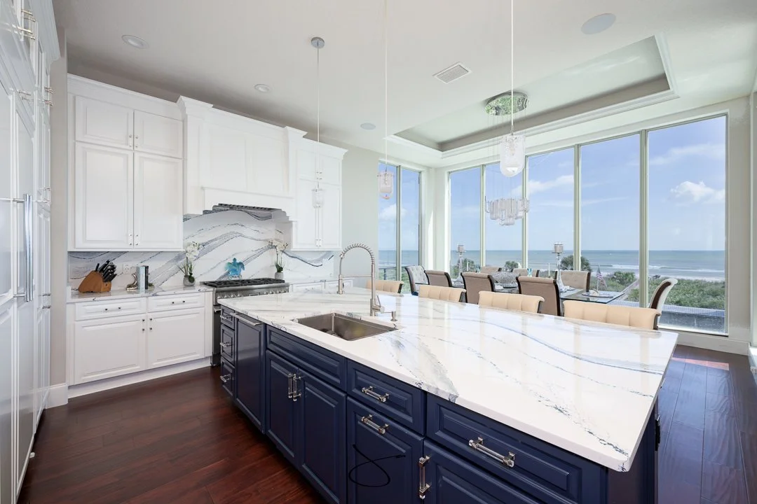 Modern kitchen with white cabinets, dark blue island, marble countertops, large windows showing ocean view, and a dining area with a chandelier.