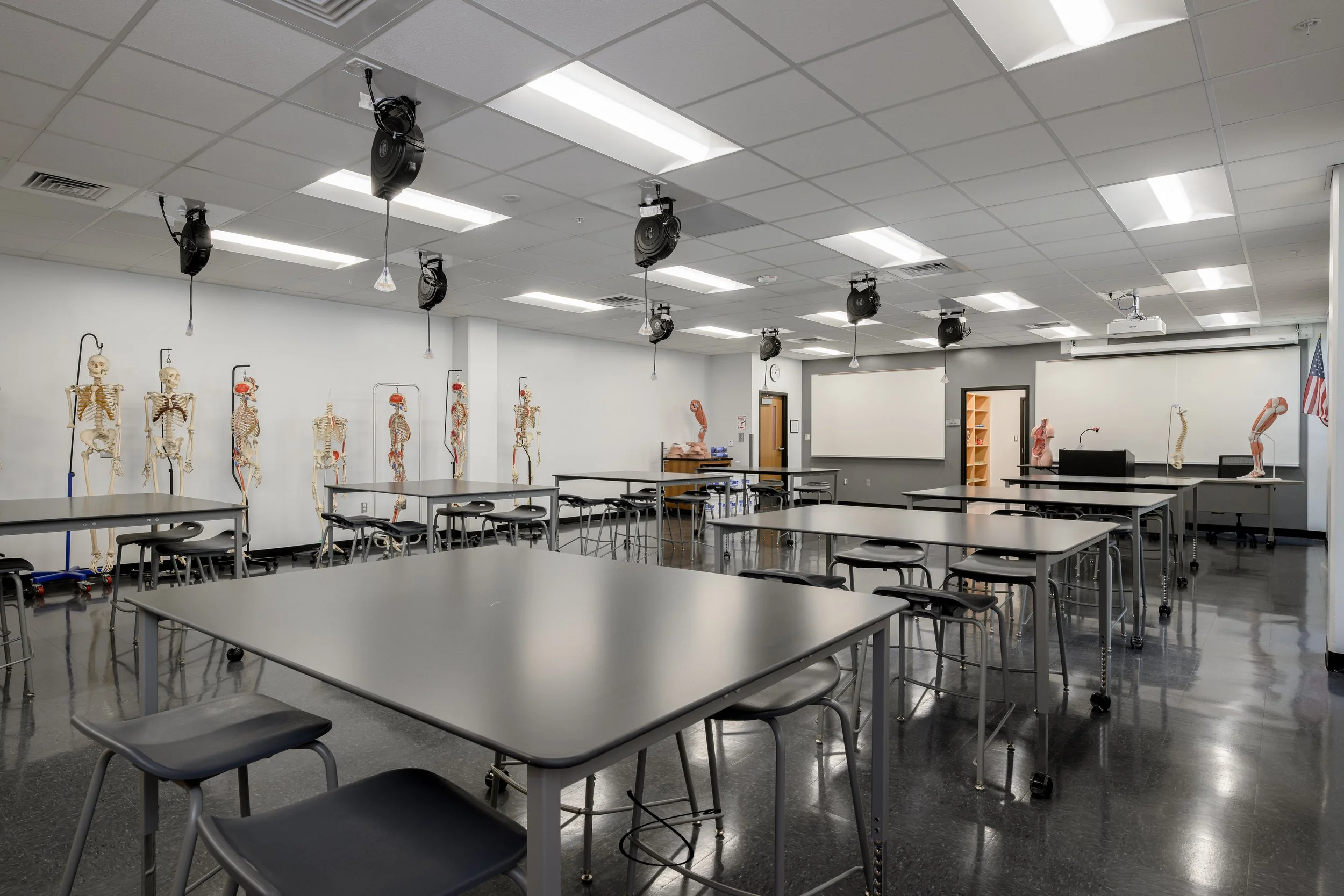 Empty science classroom or laboratory with skeleton models placed along the back wall, several tables with chairs, a projector hanging from the ceiling, and a door in the back. The room has bright white lighting and gray flooring.