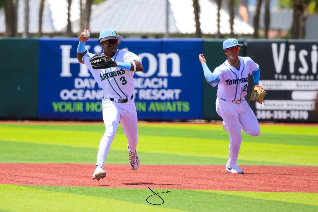 Two baseball players from the Tortugas team celebrating on the field, with one jumping and the other preparing to throw the ball, in a stadium with advertisements in the background.