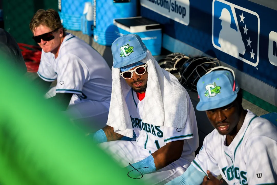 Three baseball players sitting in the dugout, wearing white jerseys with teal and green accents, and light blue caps with a logo featuring a green creature. One player has a towel over his head and is wearing sunglasses, while the others are also wea