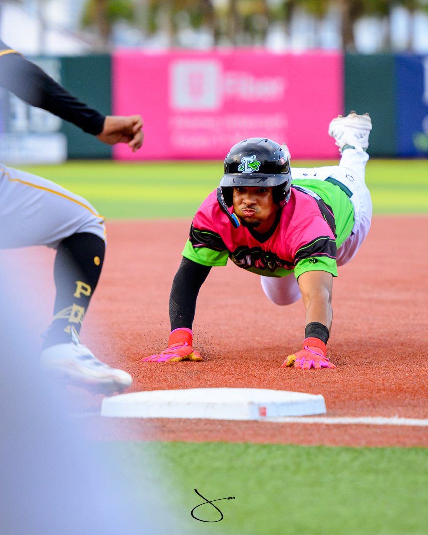 A baseball player in pink and green uniform is sliding into a base on the field, with another player approaching the base, and an umpire nearby.