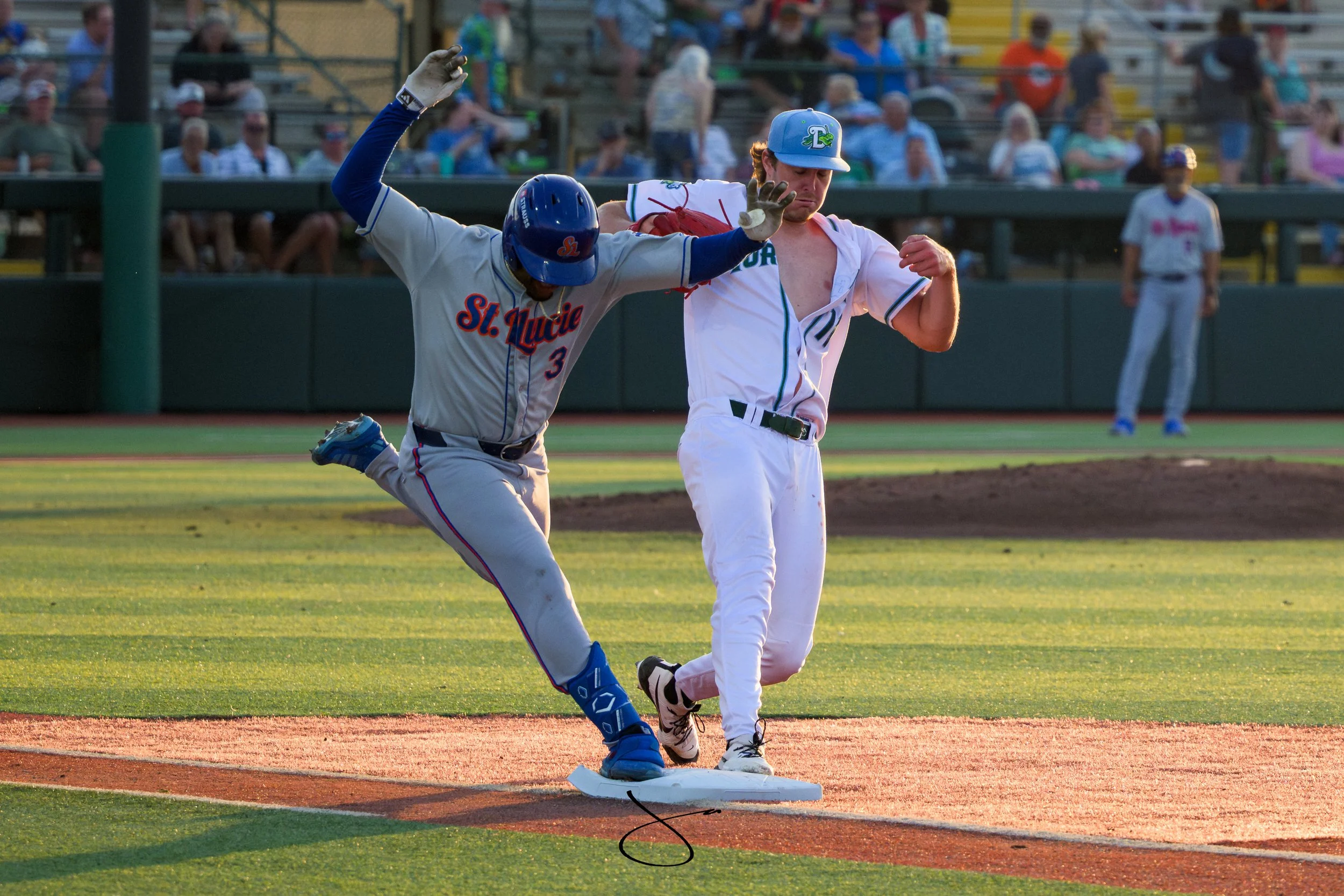 A baseball player from St. Lucie running on a base while another player from the opposing team steps on the base to tag him out during a game, with spectators watching in the background.