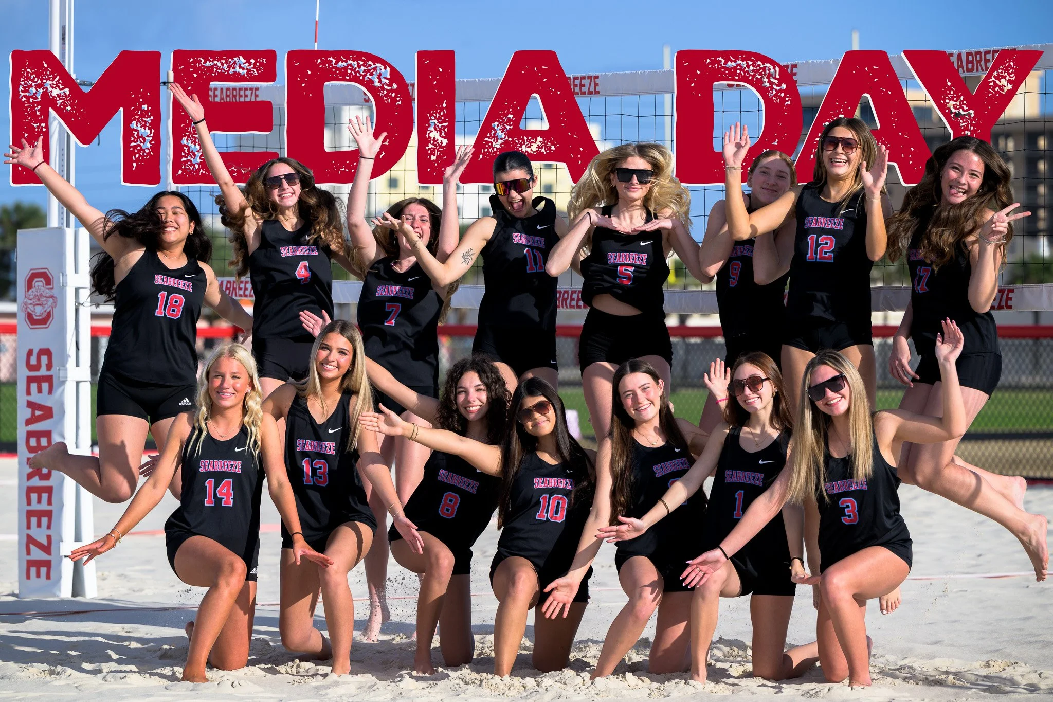 Group of young women in volleyball uniforms celebrating on a beach volleyball court with a large sign reading 'MEDA DAY' in the background.