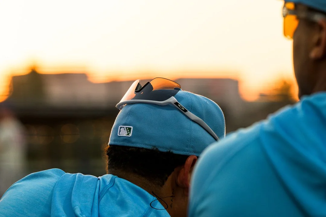 Close-up of two baseball players in blue uniforms, with the back of one player's head and the other player's shoulder visible, at sunset.