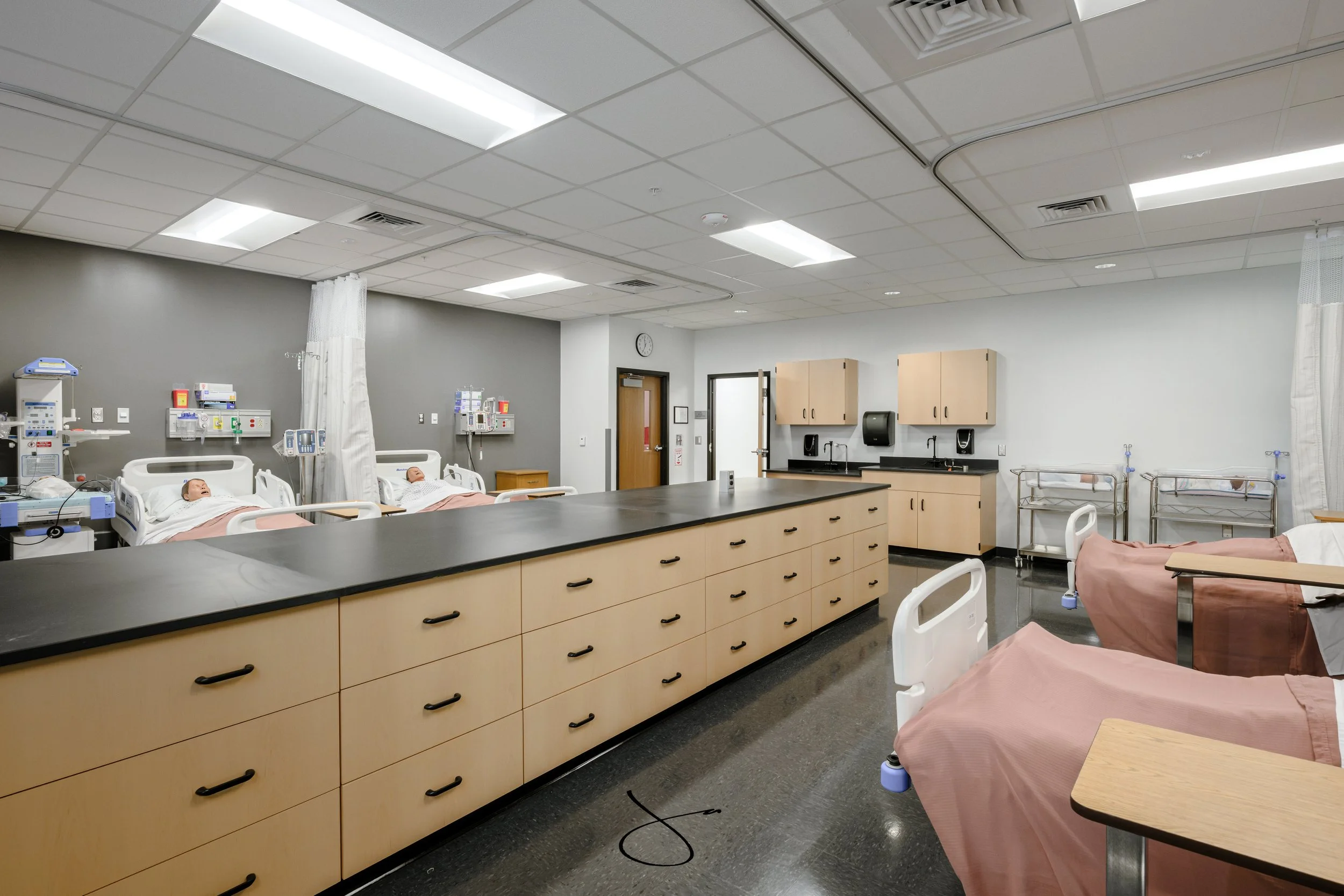 A hospital room with multiple beds, medical equipment, and medical mannequins simulating patients. The room has a ceiling with fluorescent lights, cabinets, and counters, with privacy curtains and sinks for medical use.