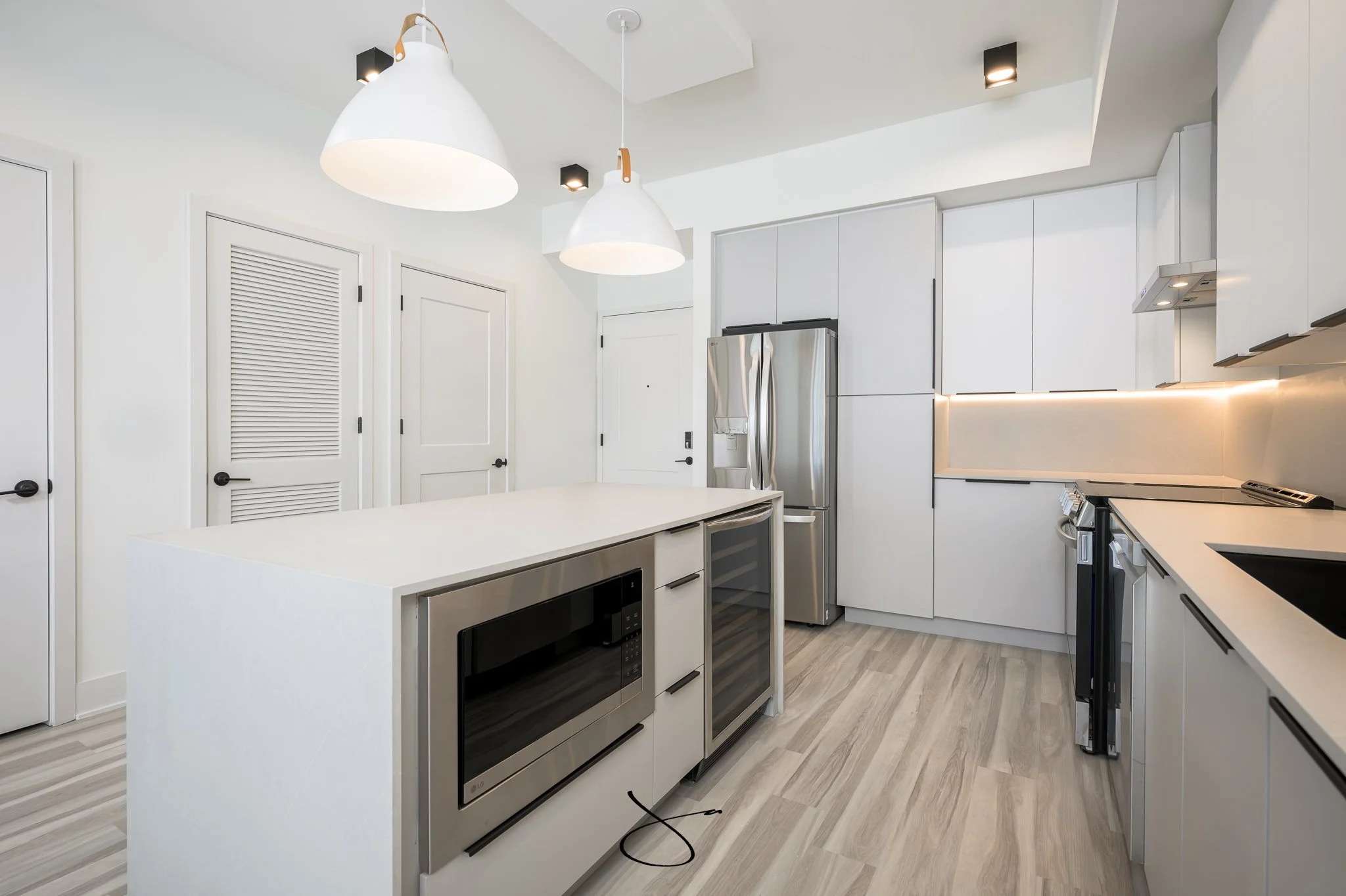 Modern white kitchen with pendant lights, stainless steel refrigerator, black and white appliances, and light wooden flooring.