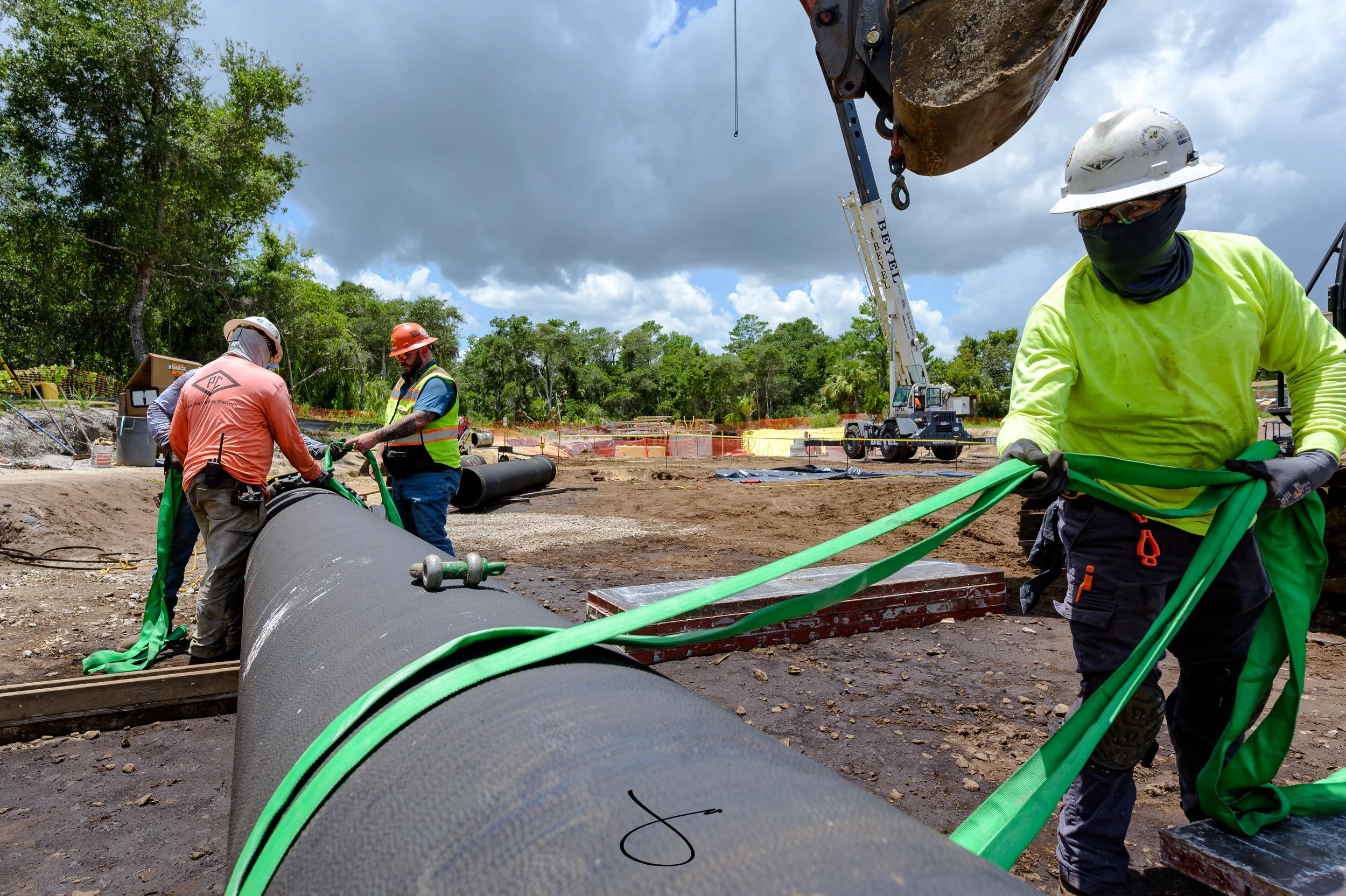 Construction workers installing or repairing large pipes in a construction site with trees and a crane in the background.