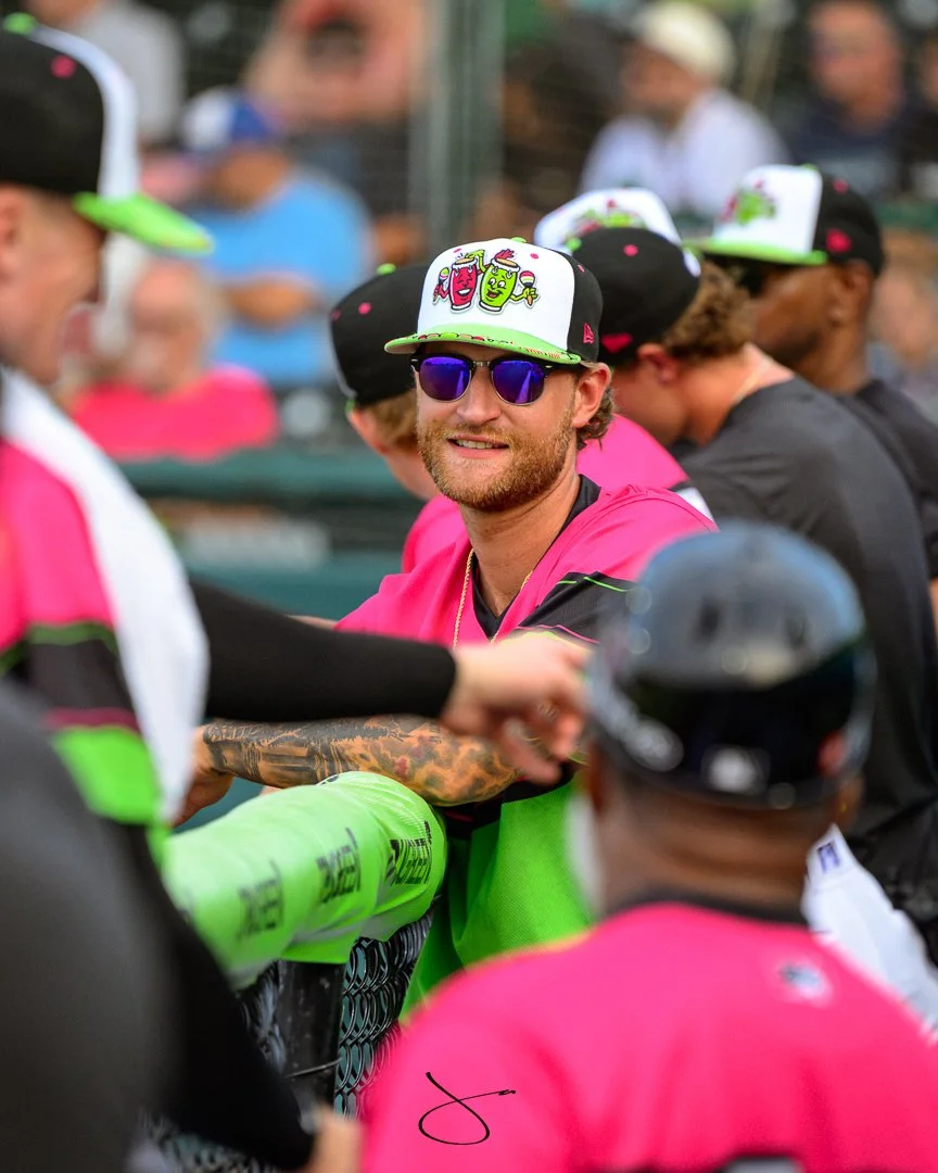 A group of baseball players in colorful uniforms and caps, with one player wearing reflective sunglasses, standing around a dugout during a game.