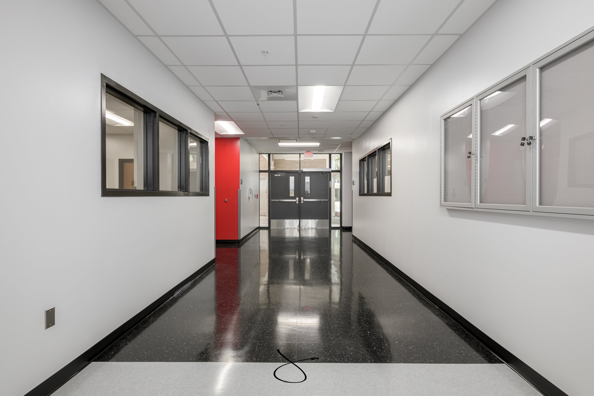 Empty college corridor with white walls, black and gray flooring, windows, and a set of glass double doors at the end.