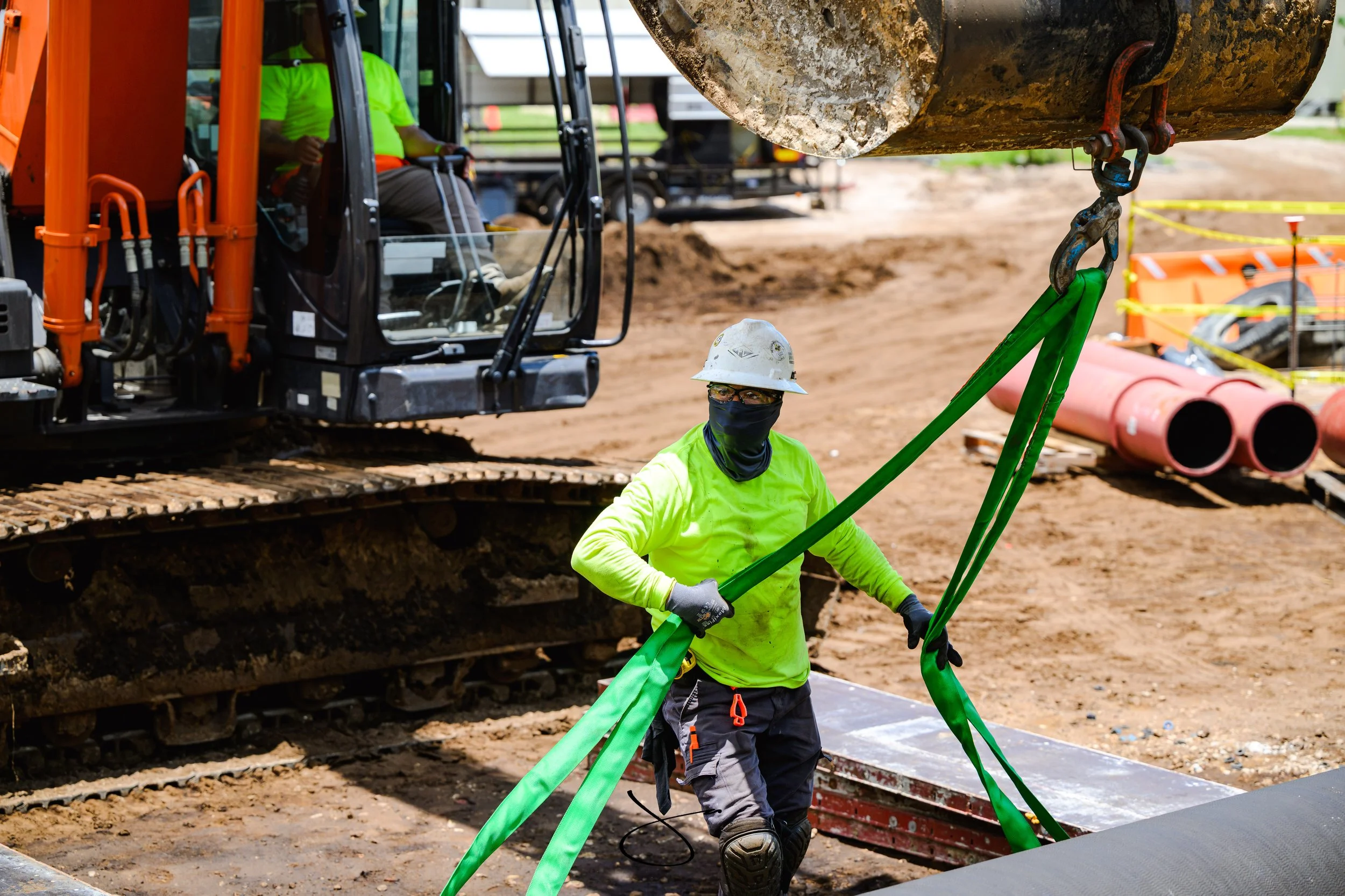 Construction worker and operator are lifting and securing a large piece of equipment using green straps and hooks on a construction site with heavy machinery and pipes in the background.