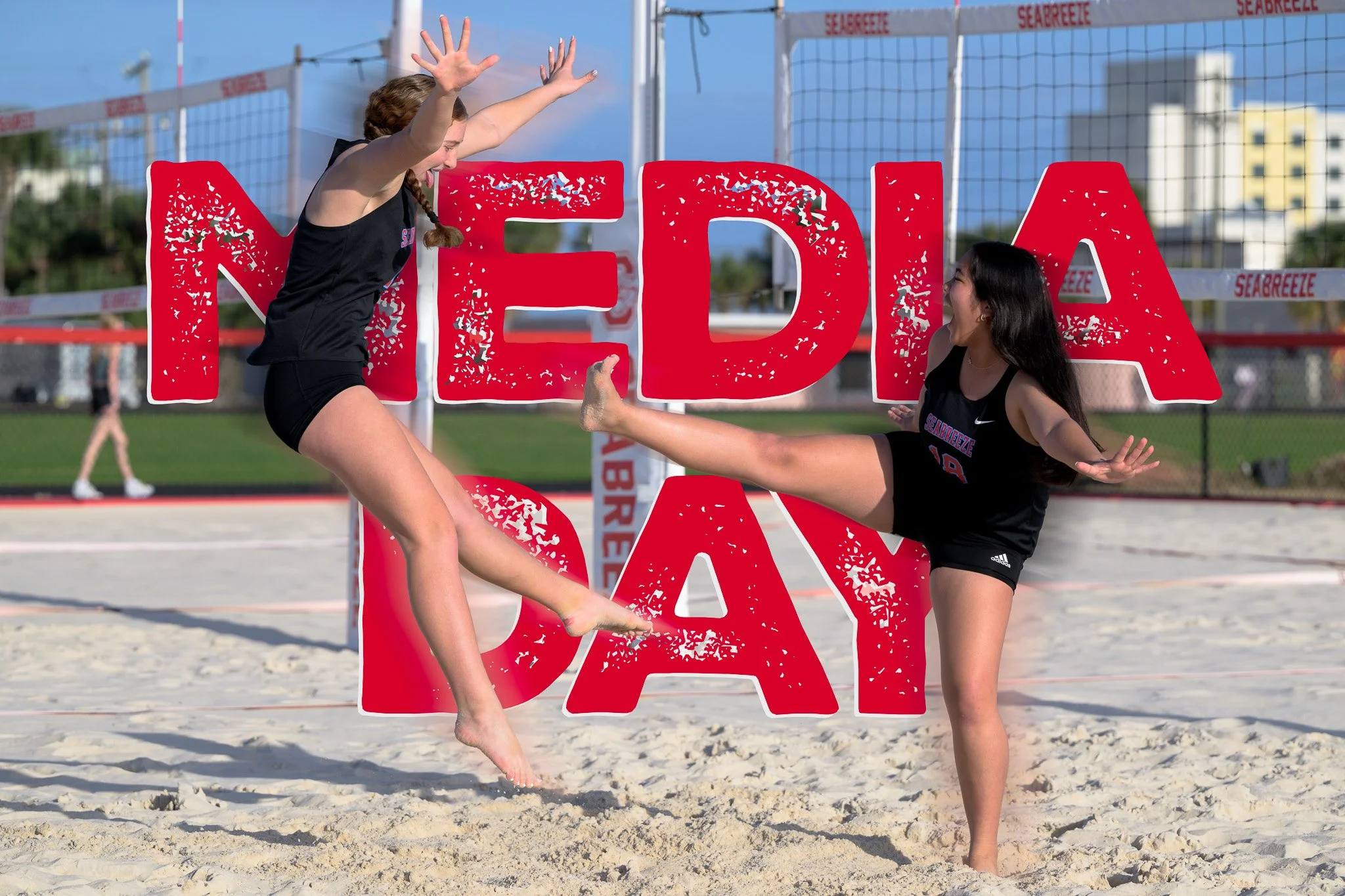 Two young girls practicing beach volleyball on a sand court, with a large red sign that reads 'NEDIA DAY' in the background.