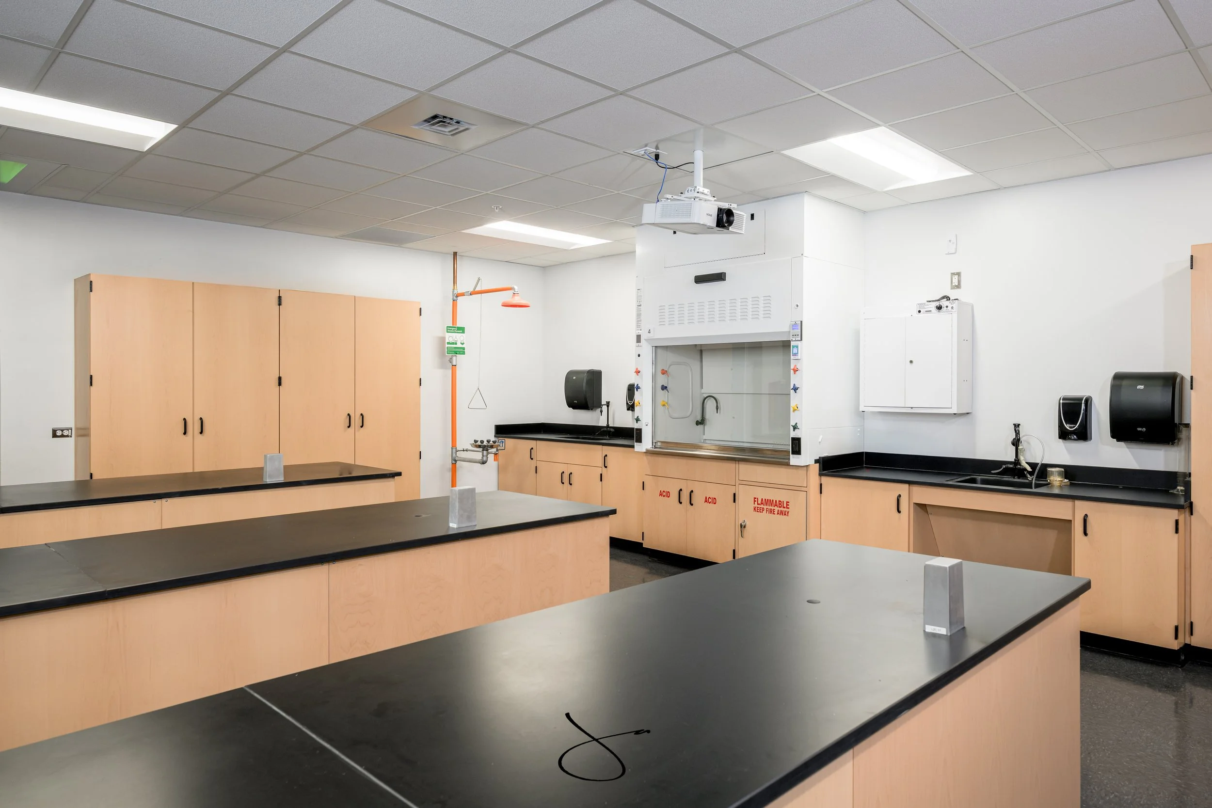 Interior view of a science classroom with black lab tables, cabinets, a fume hood, and a ceiling mounted projector.