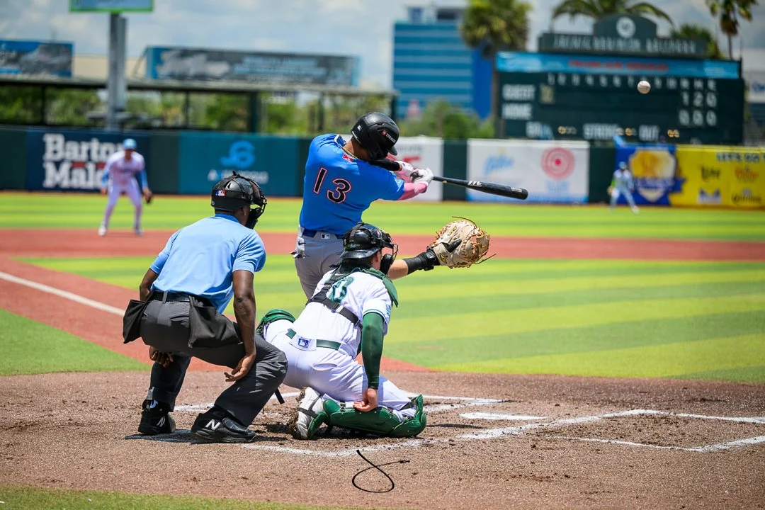 A baseball player in a blue jersey with the number 13 is swinging a bat at a pitched ball. The catcher in white and green gear is reaching out with his glove, and an umpire in a light blue shirt watches closely. Two other players are visible in the b