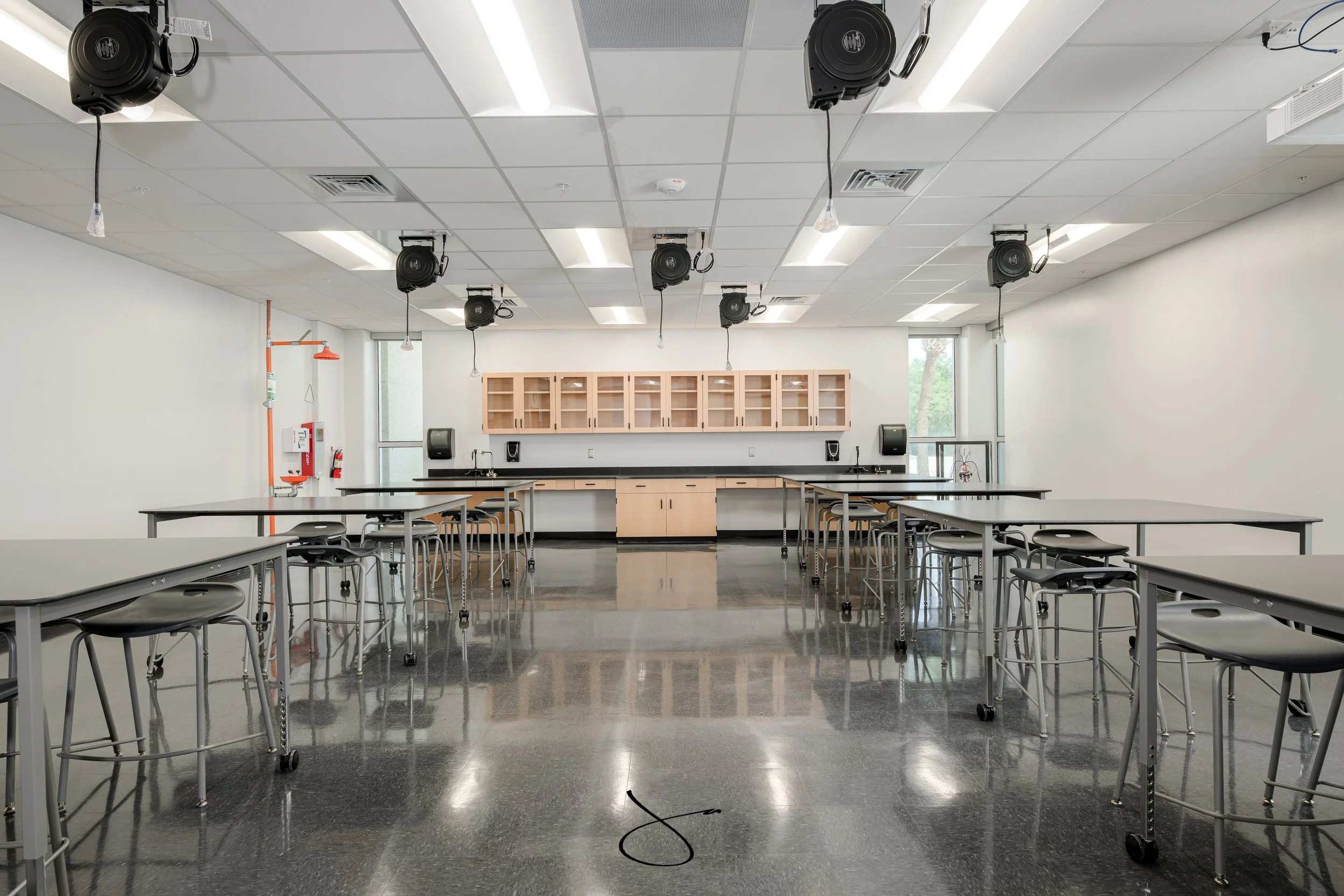 Empty classroom or training room with tables and chairs, ceiling-mounted speakers, cabinets, and windows, with cords hanging from ceiling fixtures.