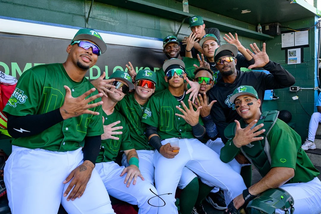 A group of young male baseball players in green and black jerseys and caps, some wearing sunglasses, posing together in a dugout.