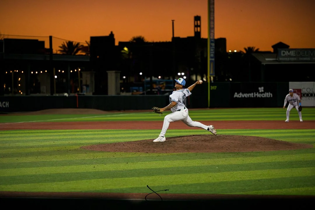A baseball player in a white uniform and cap pitching on a baseball field during sunset, with another player positioned in the background and a cityscape skyline in the distance.