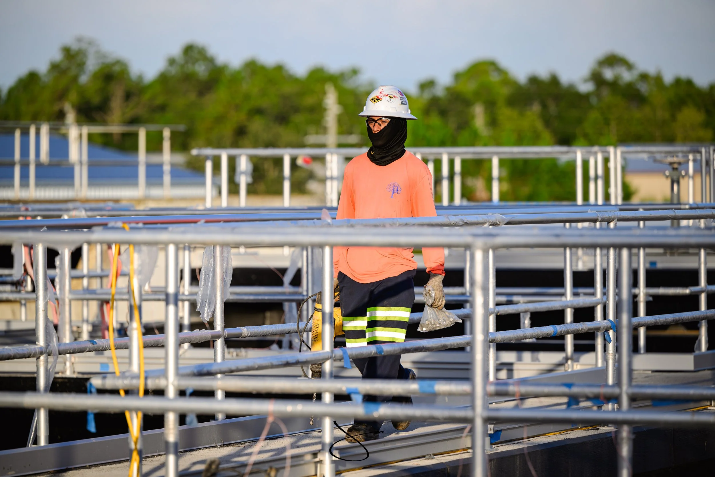 A construction worker wearing safety gear, including a white helmet, glasses, black face covering, orange shirt, and striped pants, working on a metal framework outdoors during daytime.