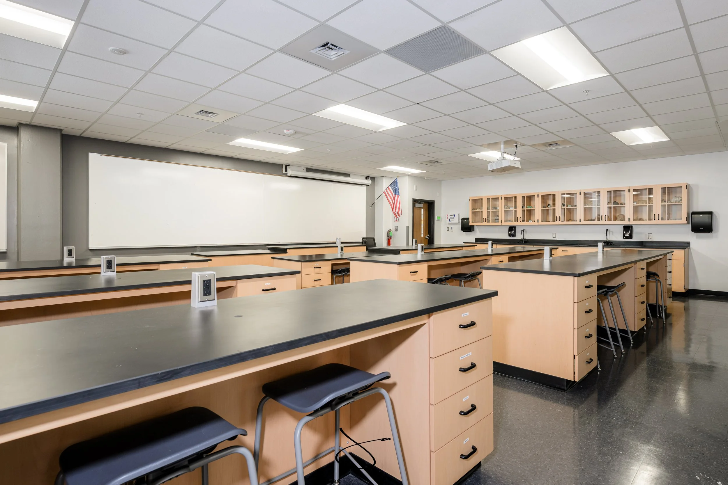 An empty classroom with black-topped tables, wooden cabinets along the back wall, and whiteboard, with American flag and classroom technology on the walls.