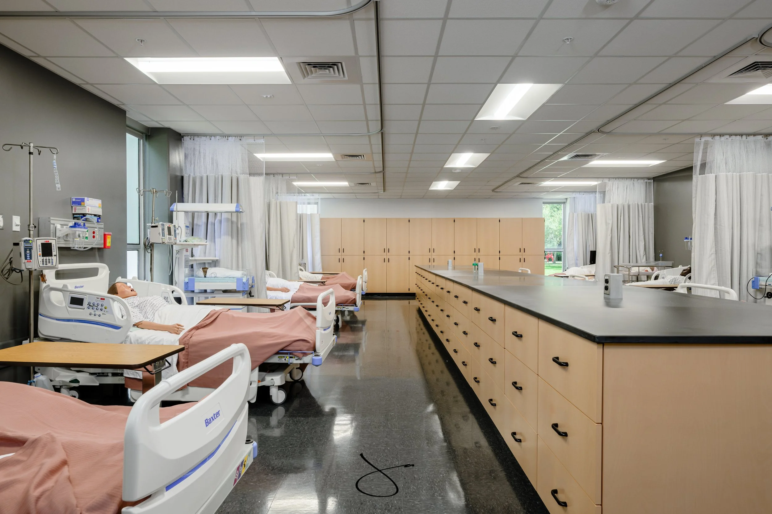 A clinical hospital room with multiple patient beds, medical equipment, and storage cabinets, illuminated by ceiling lights with windows in the background.