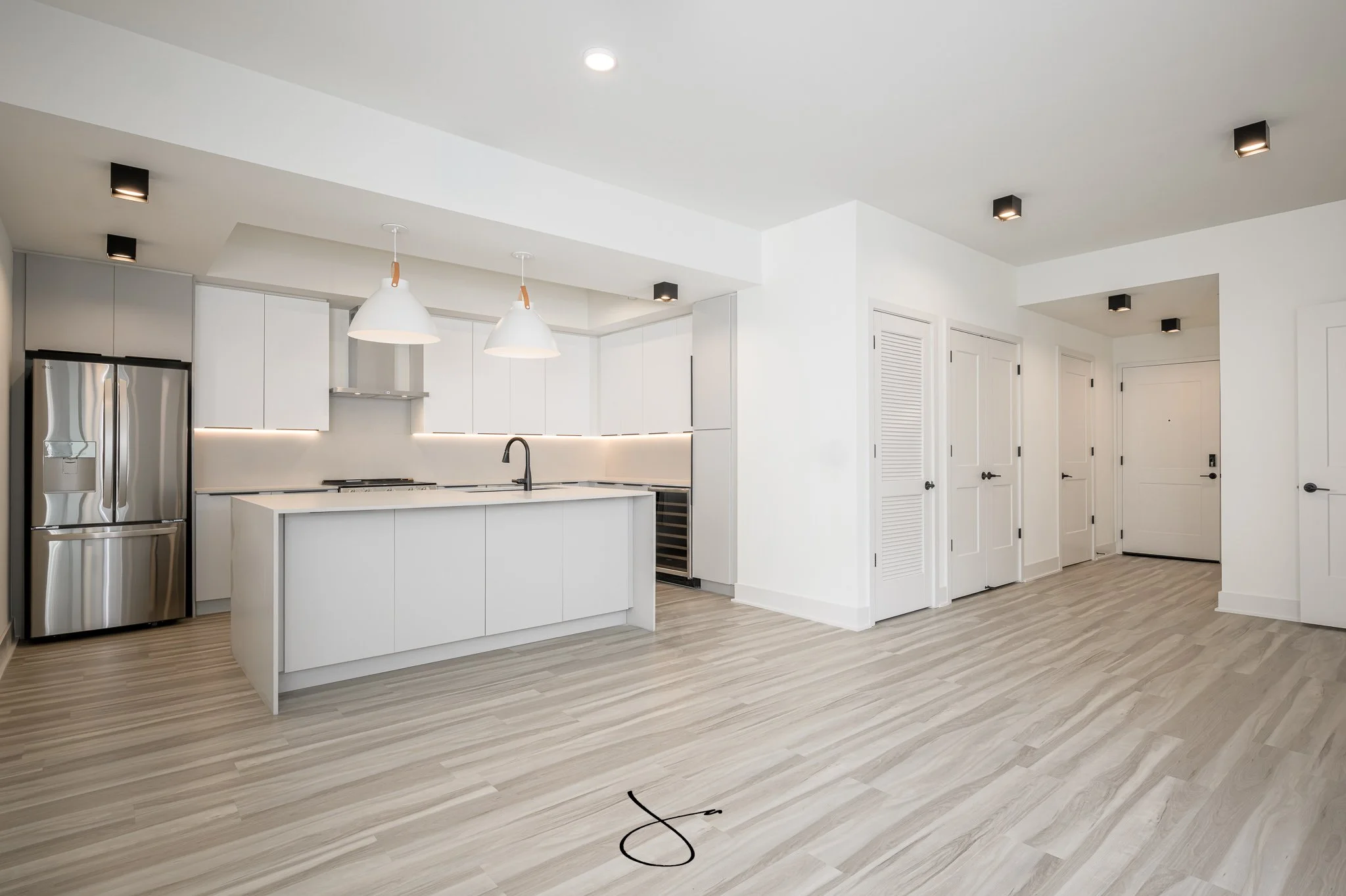 Modern white kitchen with a large island, stainless steel refrigerator, pendant lights, and wooden flooring.