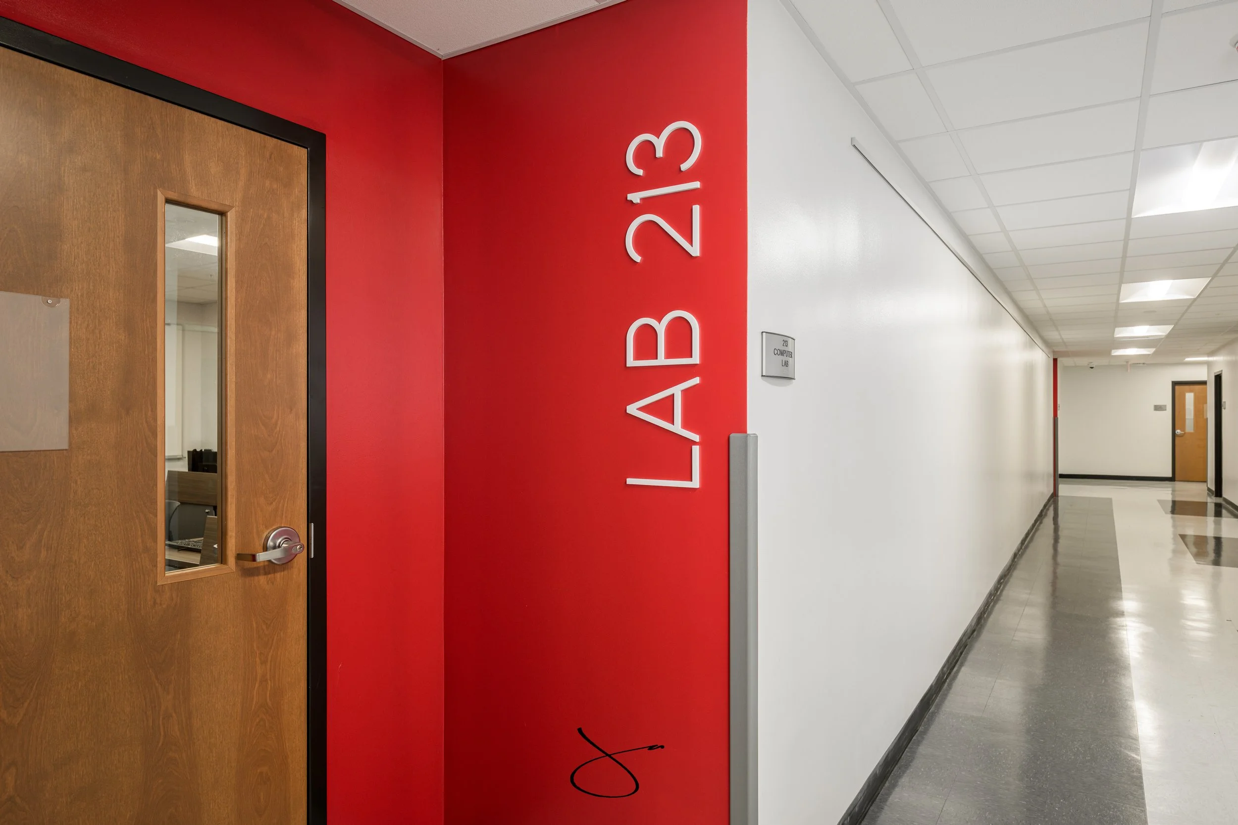 School hallway with a red wall, white lettering spelling "LAB 213," a wooden door with a small window, and a door to a computer lab at the end of the hall.