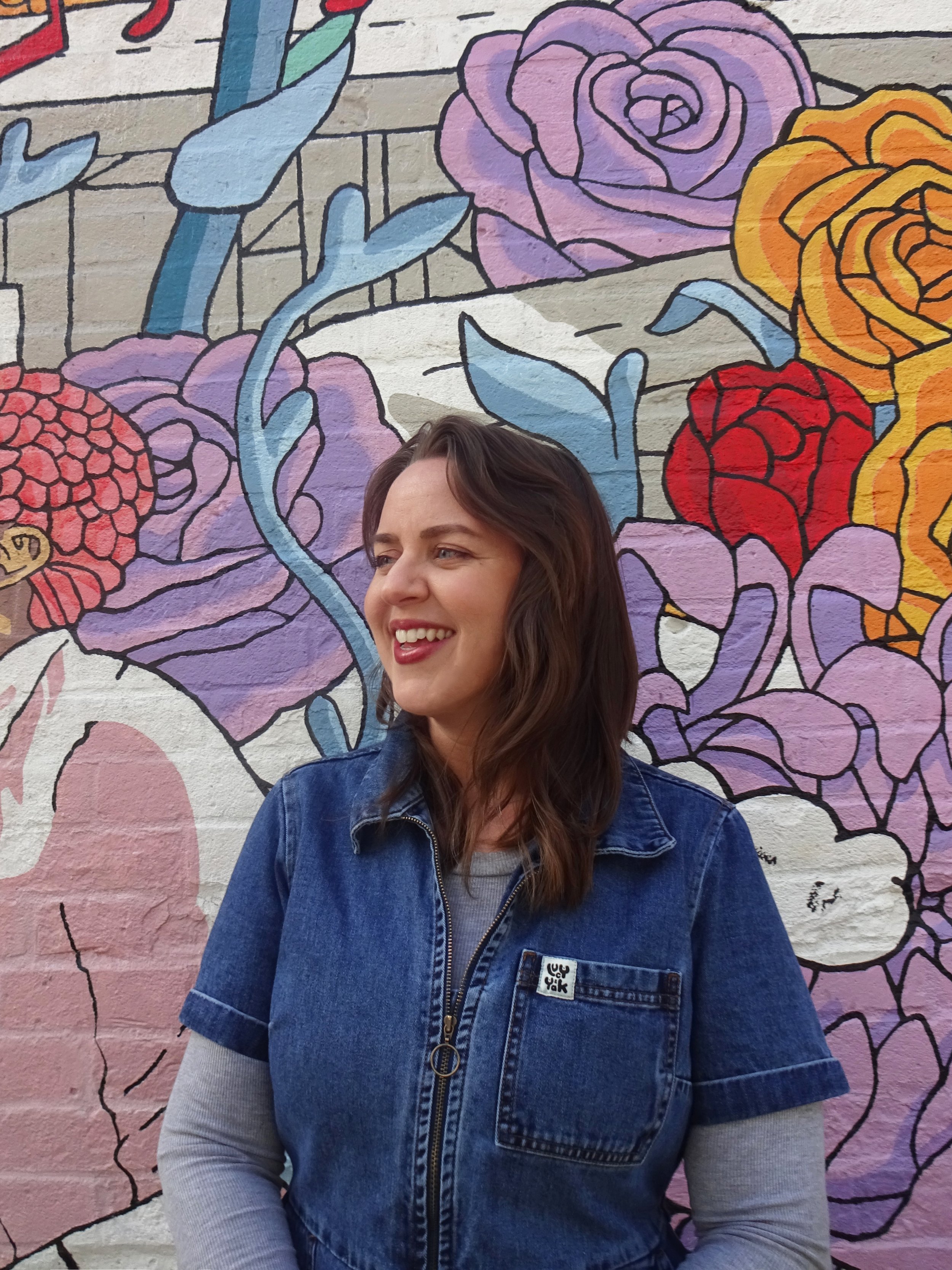 A picture of Mareika smiling and looking to the left standing in front of a floral mural