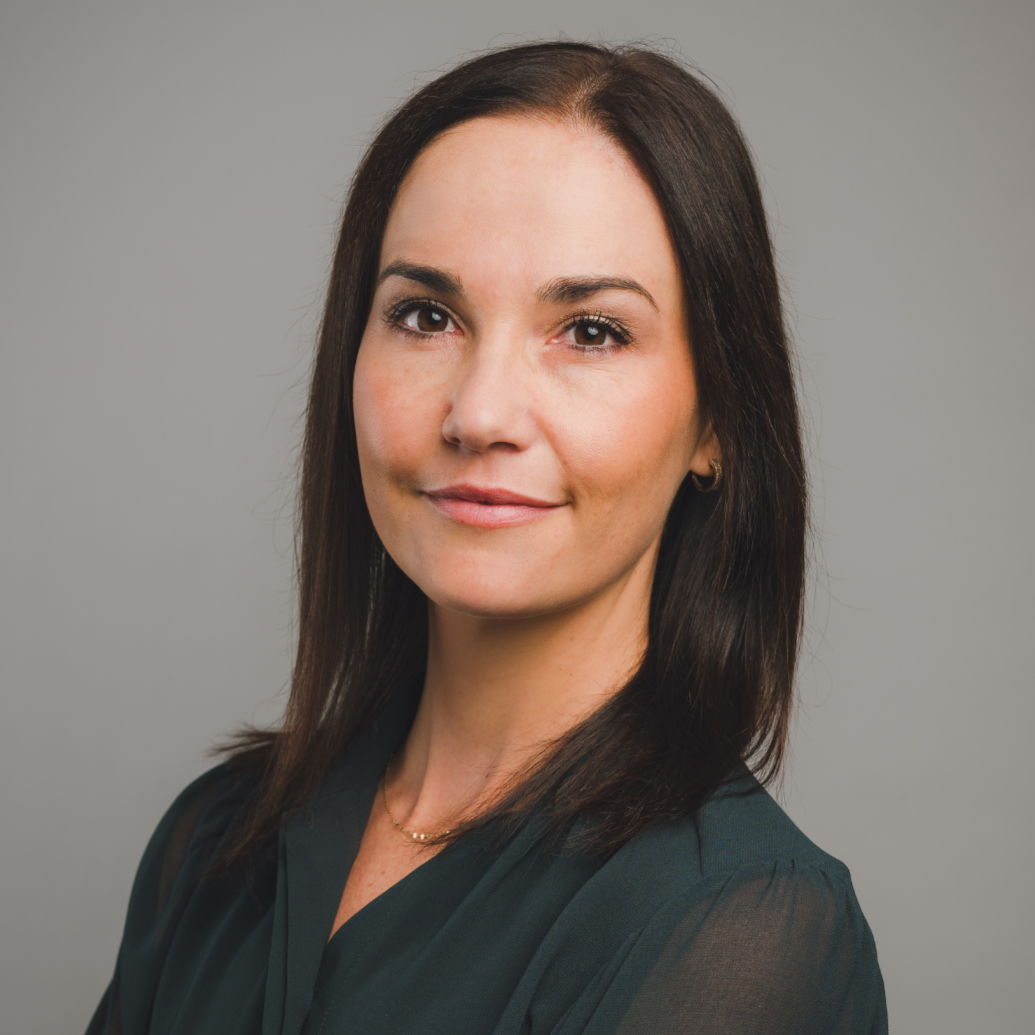 A woman with dark brown hair wearing a black blouse, posing with arms crossed against a plain gray background