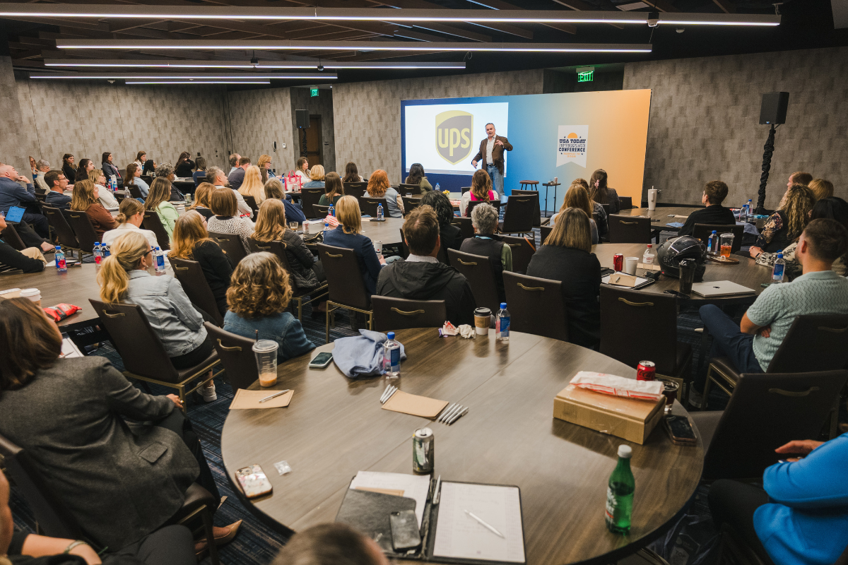 Conference room filled with attendees listening to a speaker presenting at the UPS booth during a U.S. Today Conference.