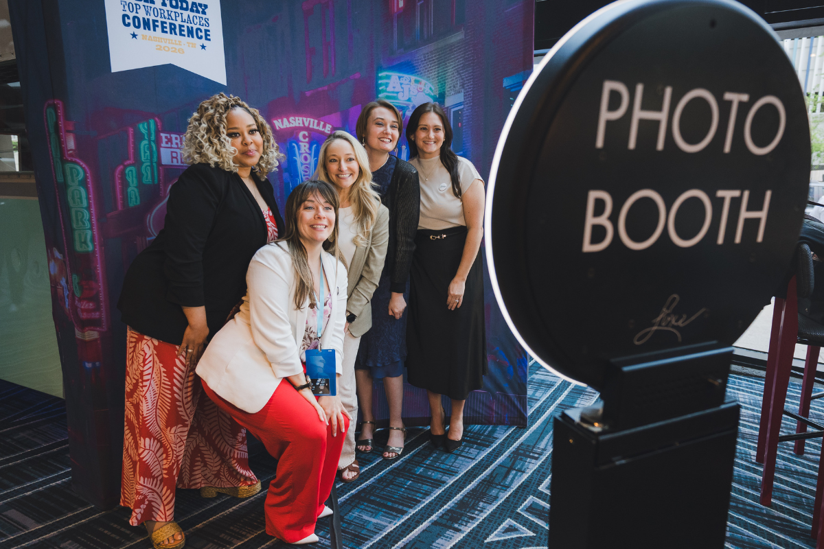 Group of five women posing in front of a colorful neon cityscape background at a conference, with a sign indicating it's a top workplaces conference in Nashville 2026, and a photo booth sign in the foreground.