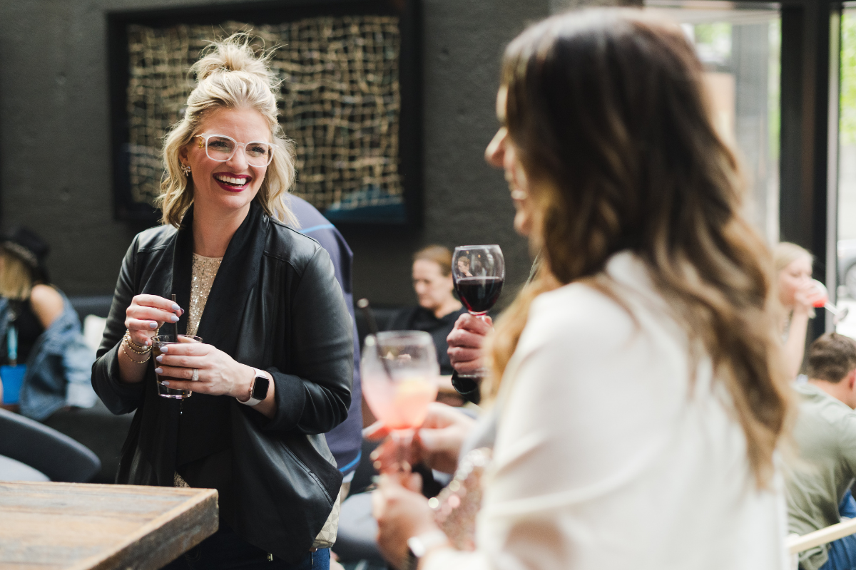 Two women socializing at a bar or cafe, holding drinks, and smiling, with other patrons in the background.