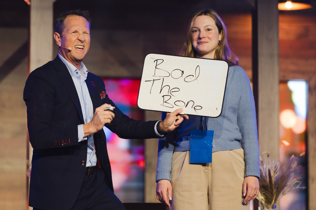 A man in a suit making a funny face holding a whiteboard with the words "Bad The Bone" written on it, standing next to a woman in a cardigan, both on stage in a venue with colorful lighting.