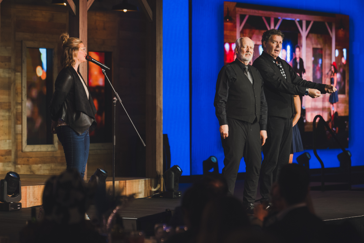 Three people on stage, two men and one woman. The woman stands at a microphone on the left, and the two men are on the right, with one of them gesturing. A large screen behind them displays a blurred image of a barn with people near it. The stage has