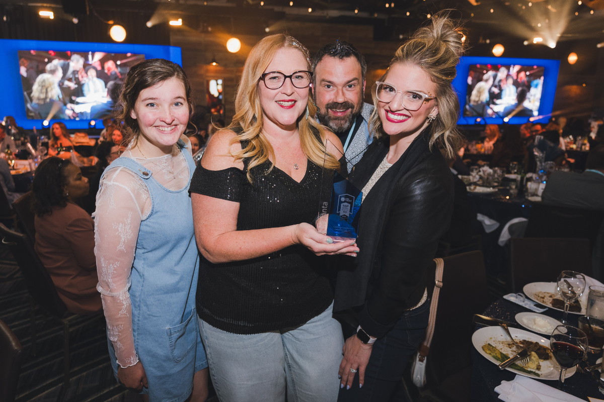 Four people, two women and a man, smiling and holding an award at a social event with many attendees, tables with food, and large screens in the background.