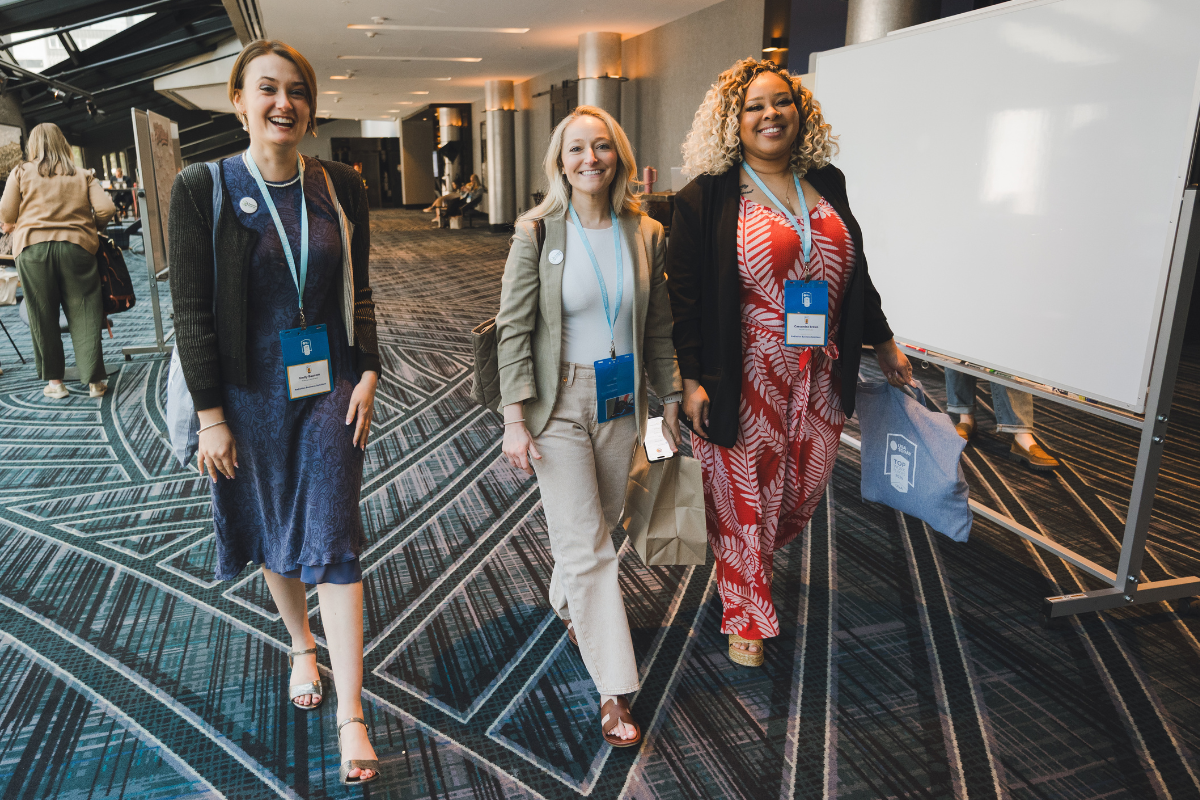 Three women walking together in a conference center, smiling, wearing badge lanyards, with a large whiteboard and other attendees in the background.
