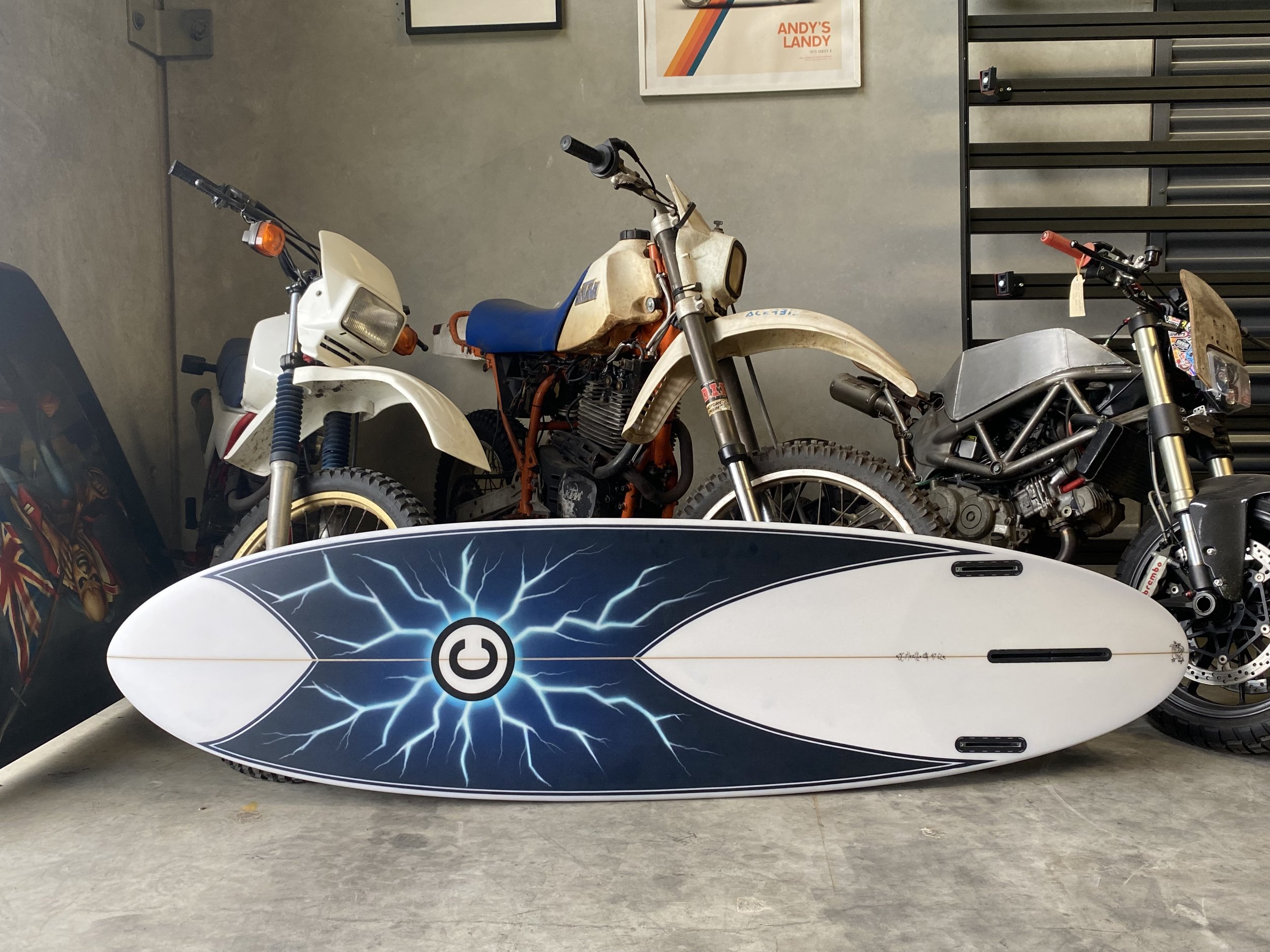 A collection of vintage motorcycles and a surfboard with lightning bolt design, displayed in a garage or workshop.