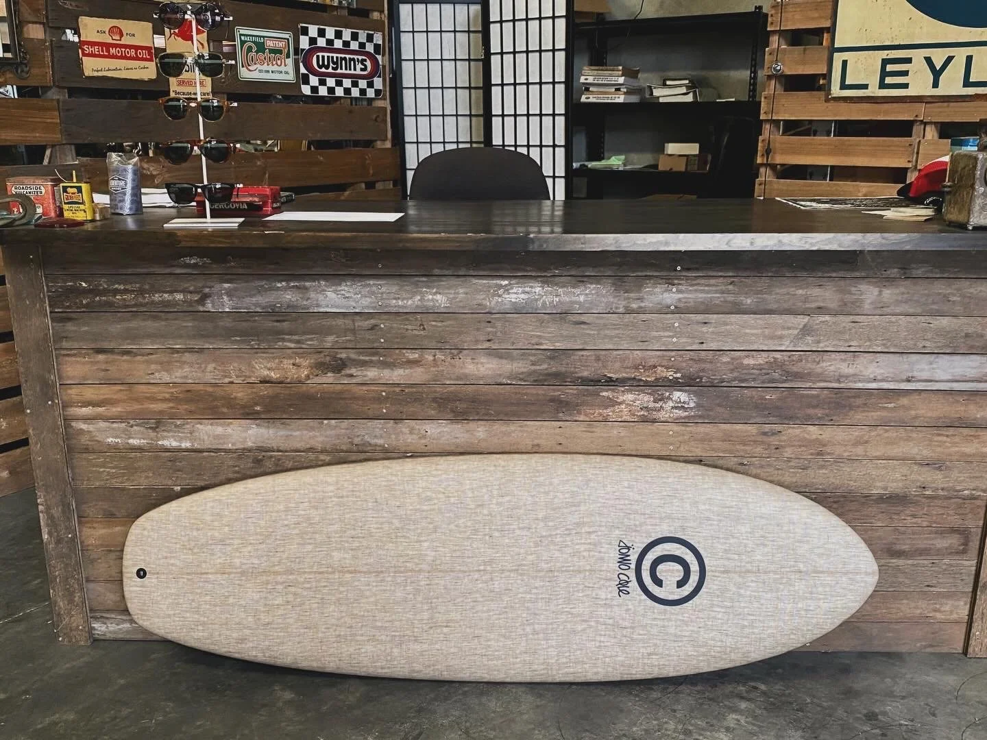 A surfboard resting against a wooden counter inside a store or workshop. The surfboard has a beige textured surface with a black logo near one end.