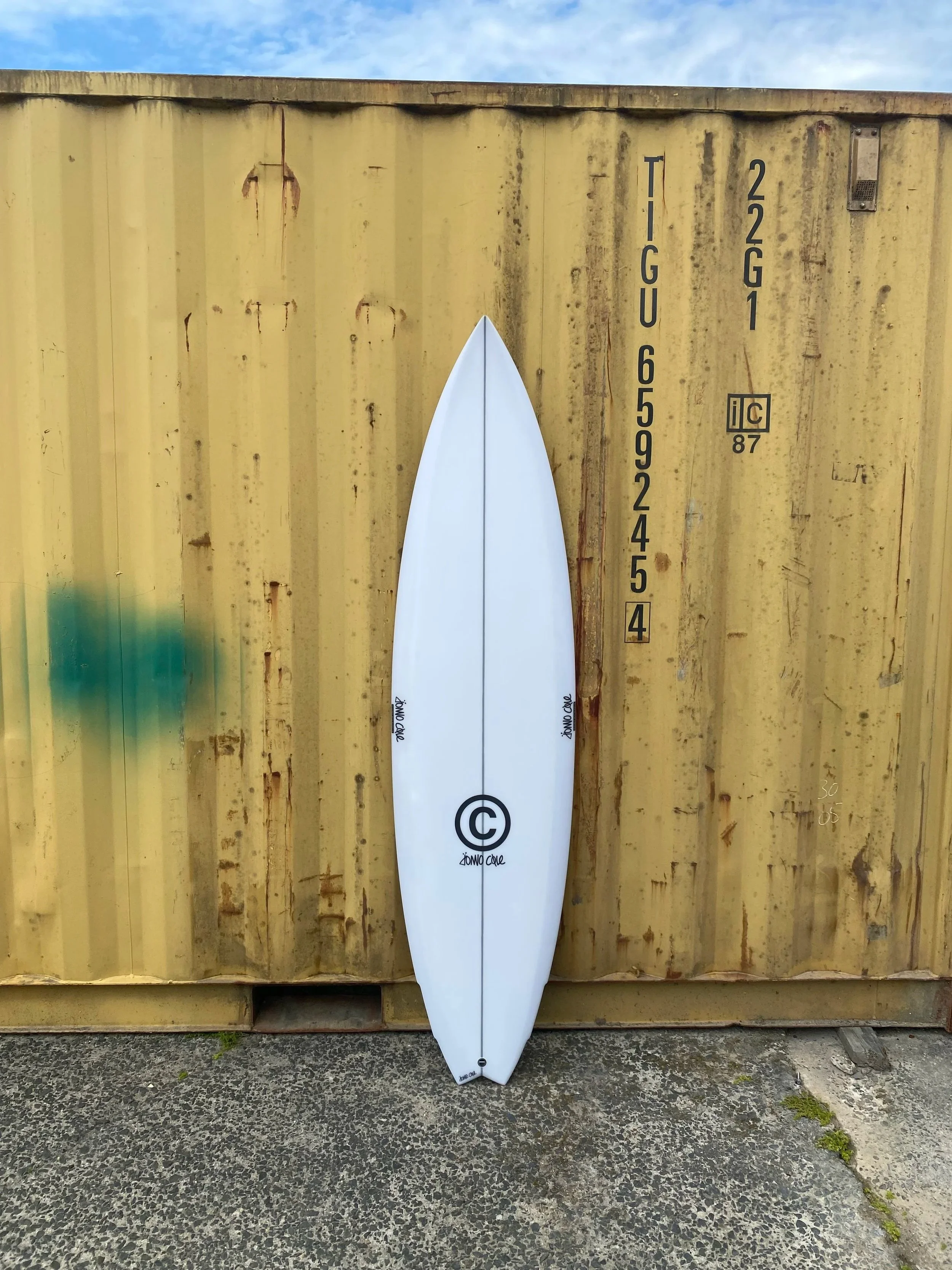 A white surfboard with black logo leaning against a yellow shipping container outdoors.