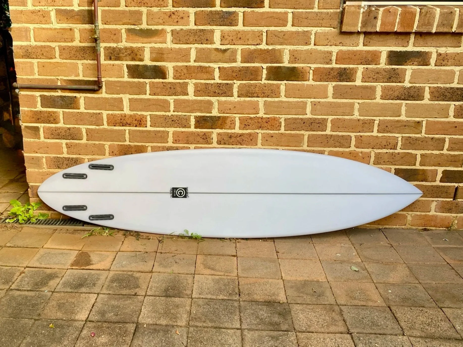A white surfboard with black traction pads, leaning against a brick wall outdoors. The surfboard is on a paved surface with some small green plants near the bottom left corner.